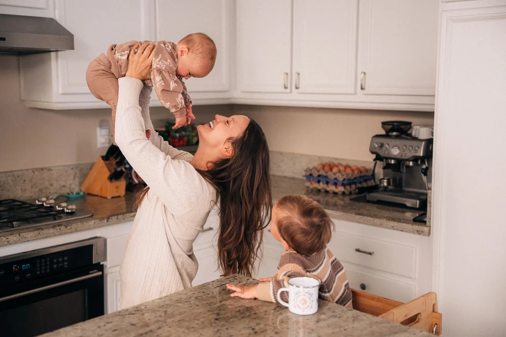 Mom lifting her baby overhead in the kitchen, mid-laugh