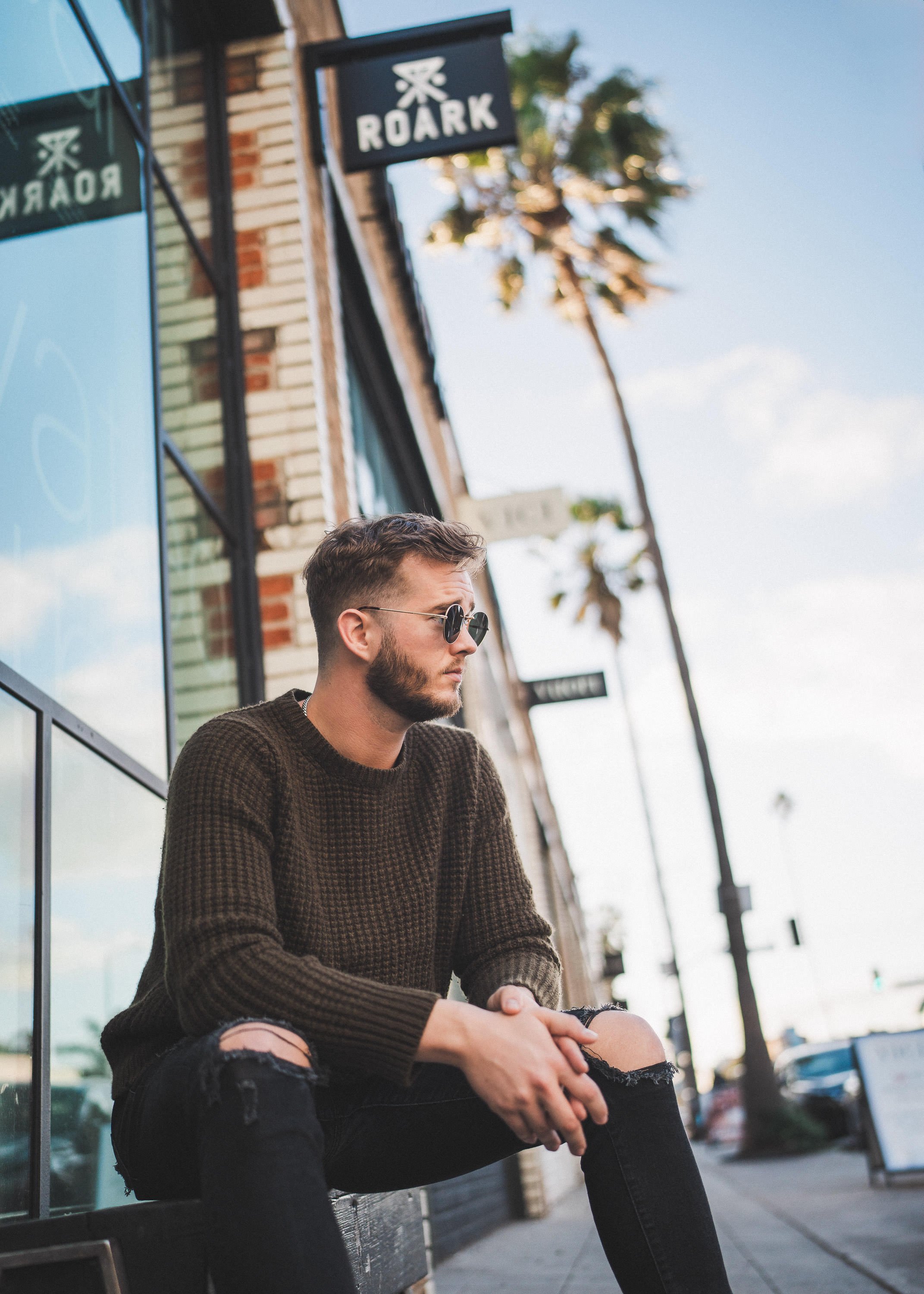 A young man with sunglasses, a brown sweater, and ripped jeans sitting on a bench outside a building with palm trees in the background.