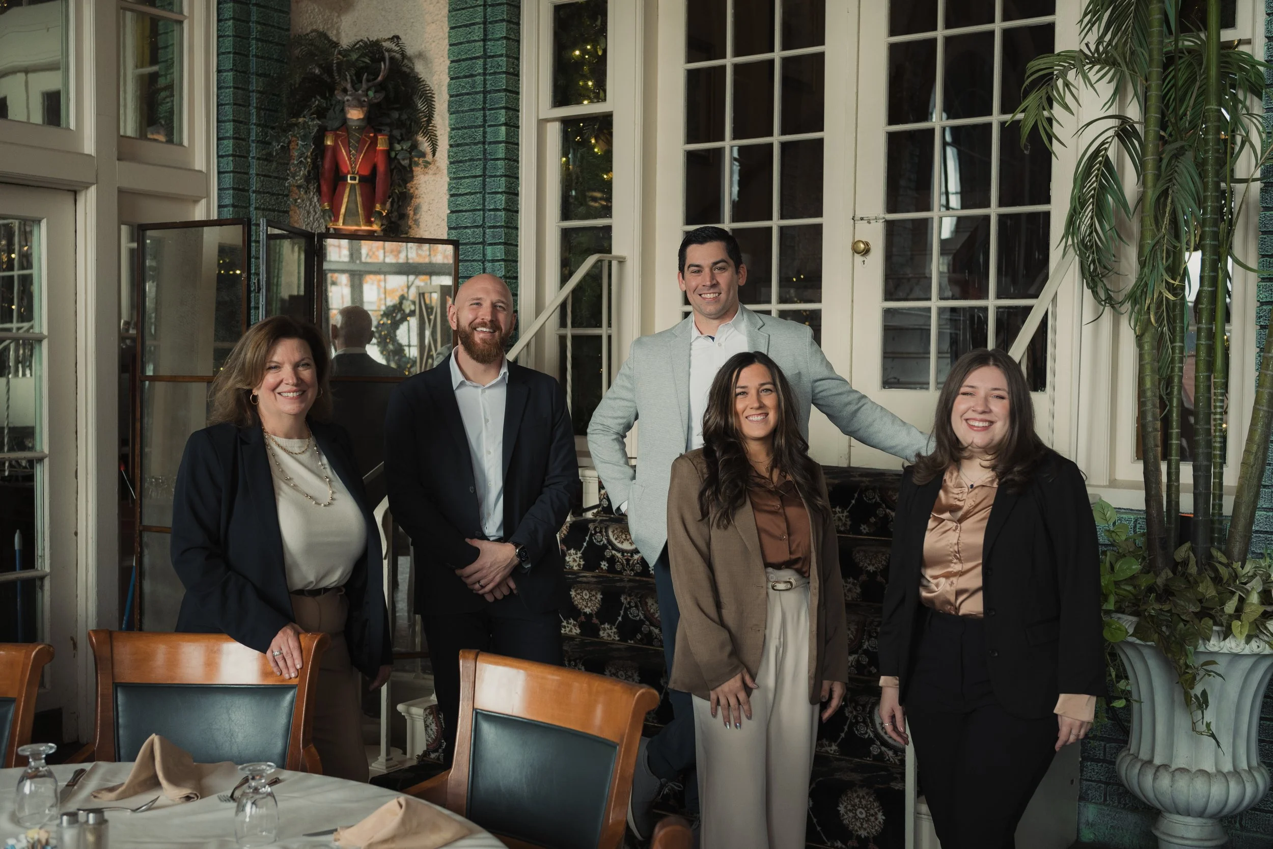 Group of five people posing indoors at a restaurant or event space, smiling at the camera, with windows, green plants, and a decorative piece in the background.