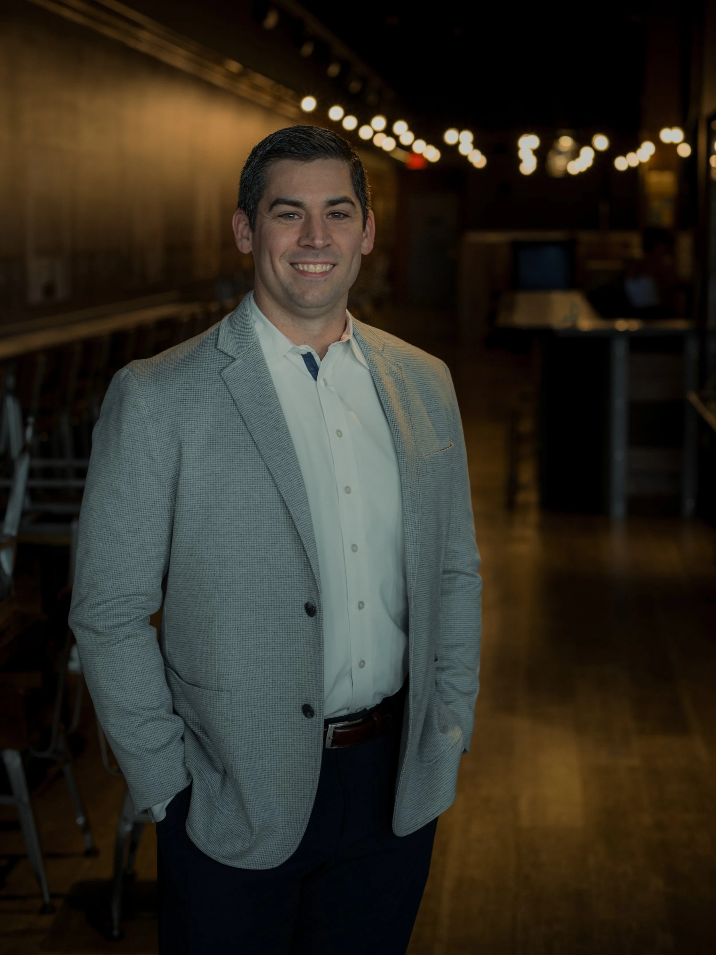 A man in a light gray blazer and white shirt standing indoors with a blurred background and string lights.