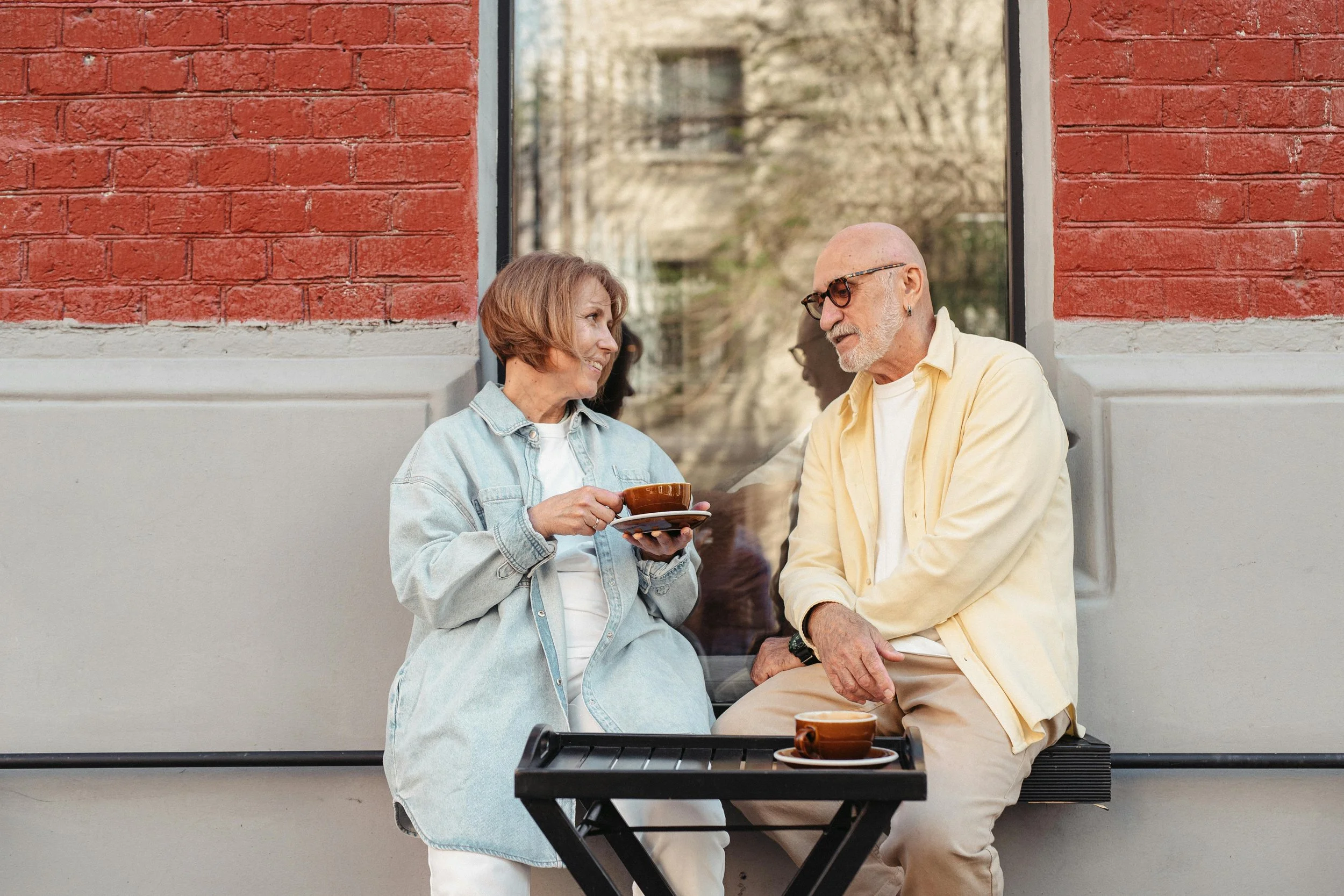 A woman and a man sit outside a cafe, smiling at each other. The woman holds a cup and saucer, and there are two cups on a small table between them. They are sitting against a large window with a brick building reflected in the glass.