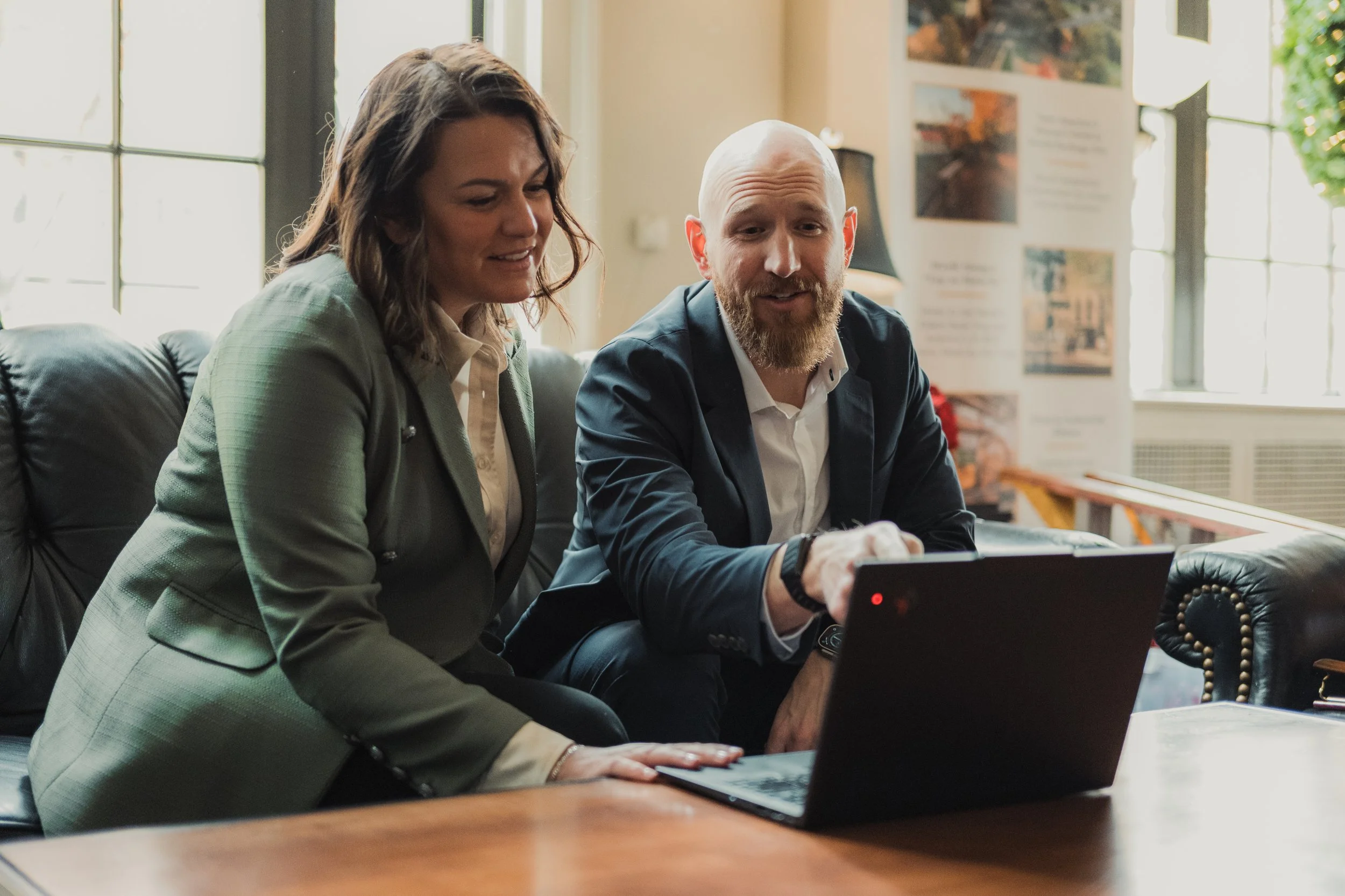 A man and woman sitting on a black leather couch looking at a laptop together in a bright room with large windows and posters on the wall.