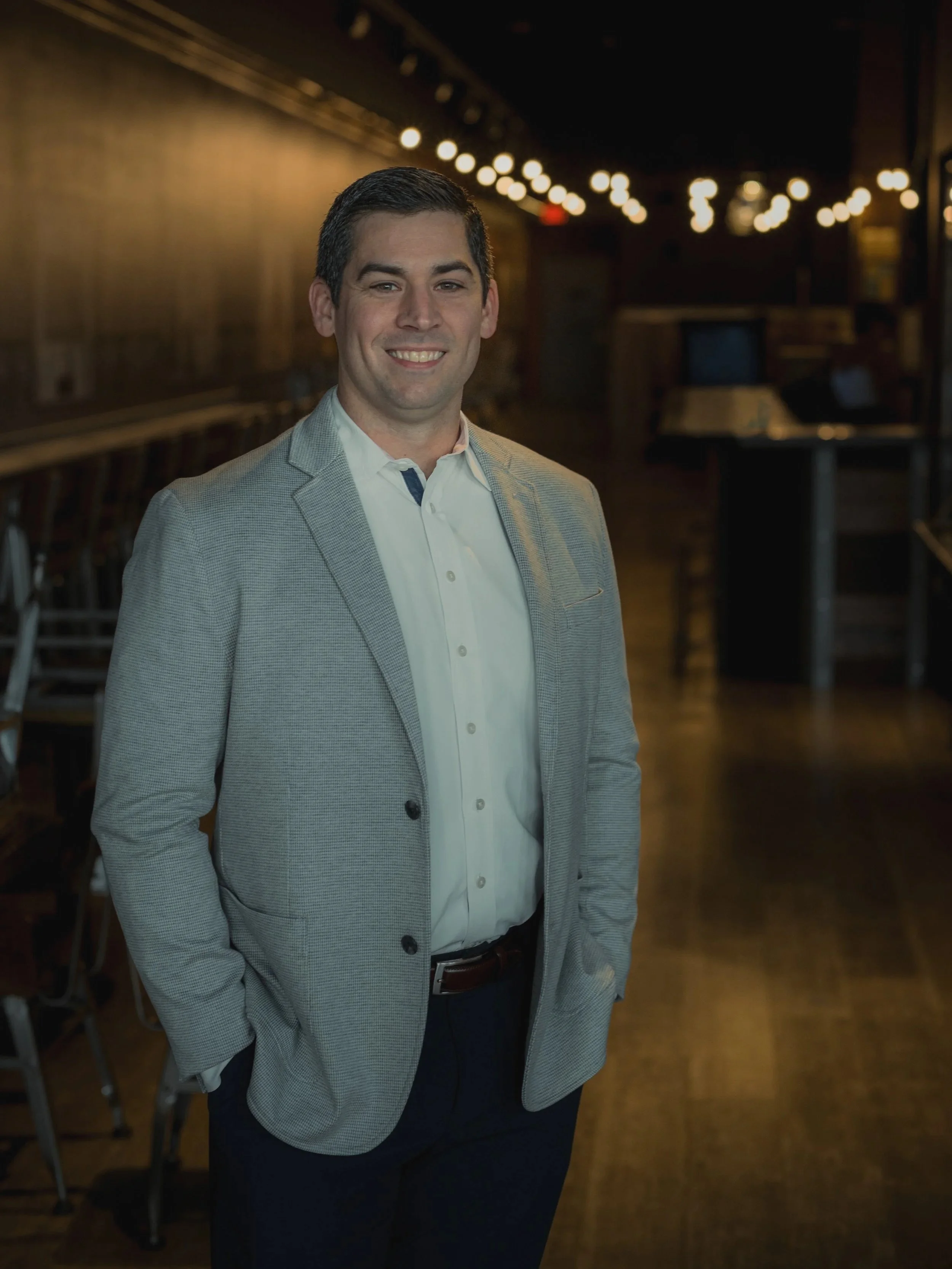 A man in a light gray blazer and white shirt standing in a dimly lit indoor space, smiling at the camera.