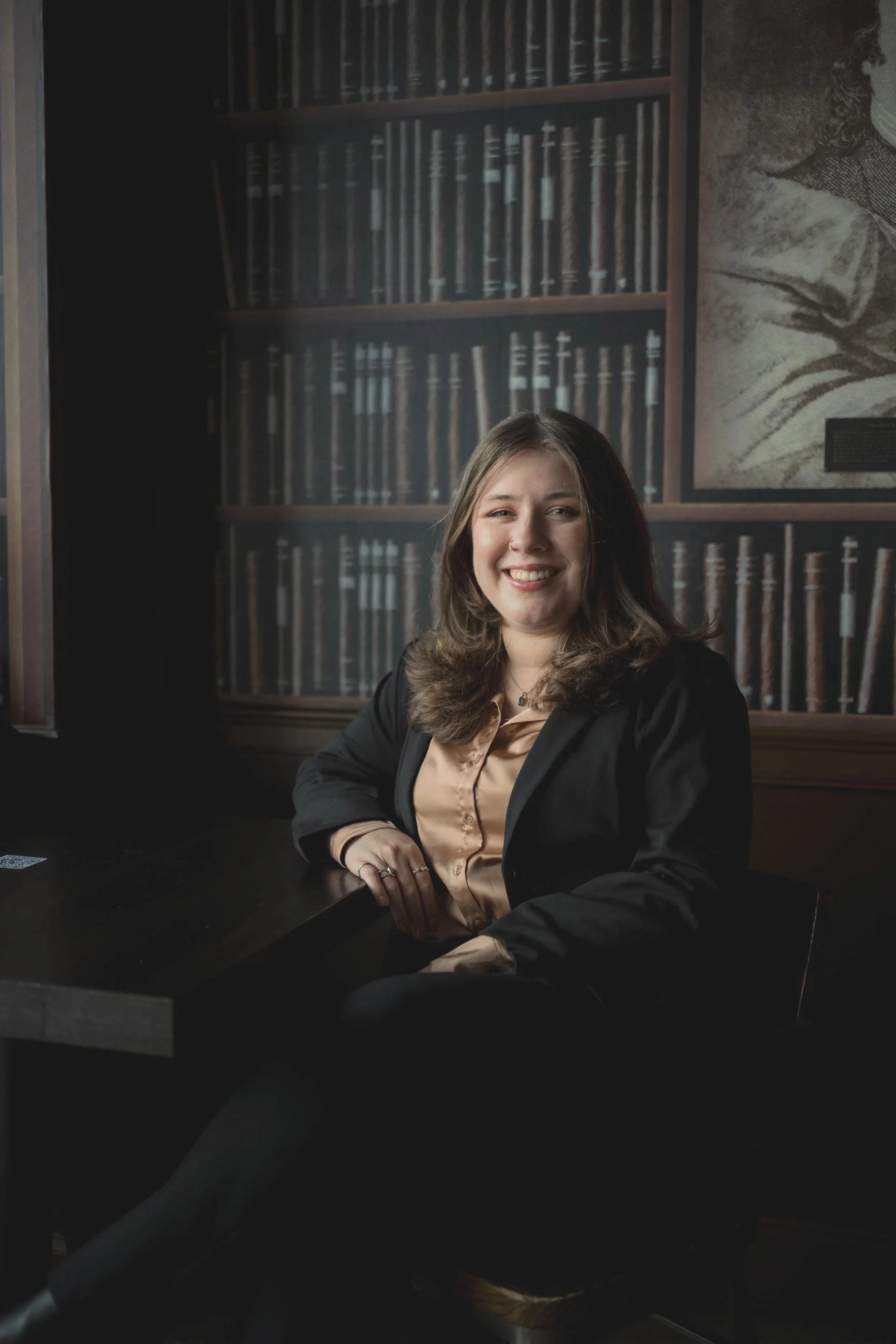 A woman seated at a table, smiling, in front of a bookshelf background.