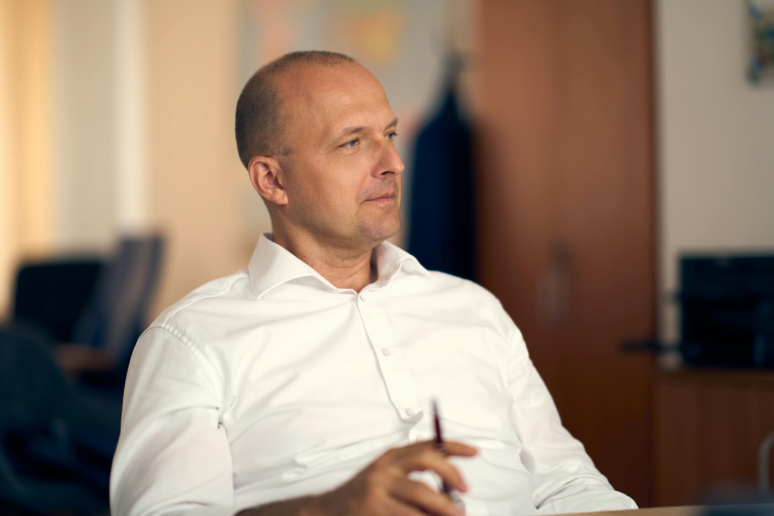 A man with a shaved head and a white dress shirt sitting in an office, holding a red pen, with a focused expression.