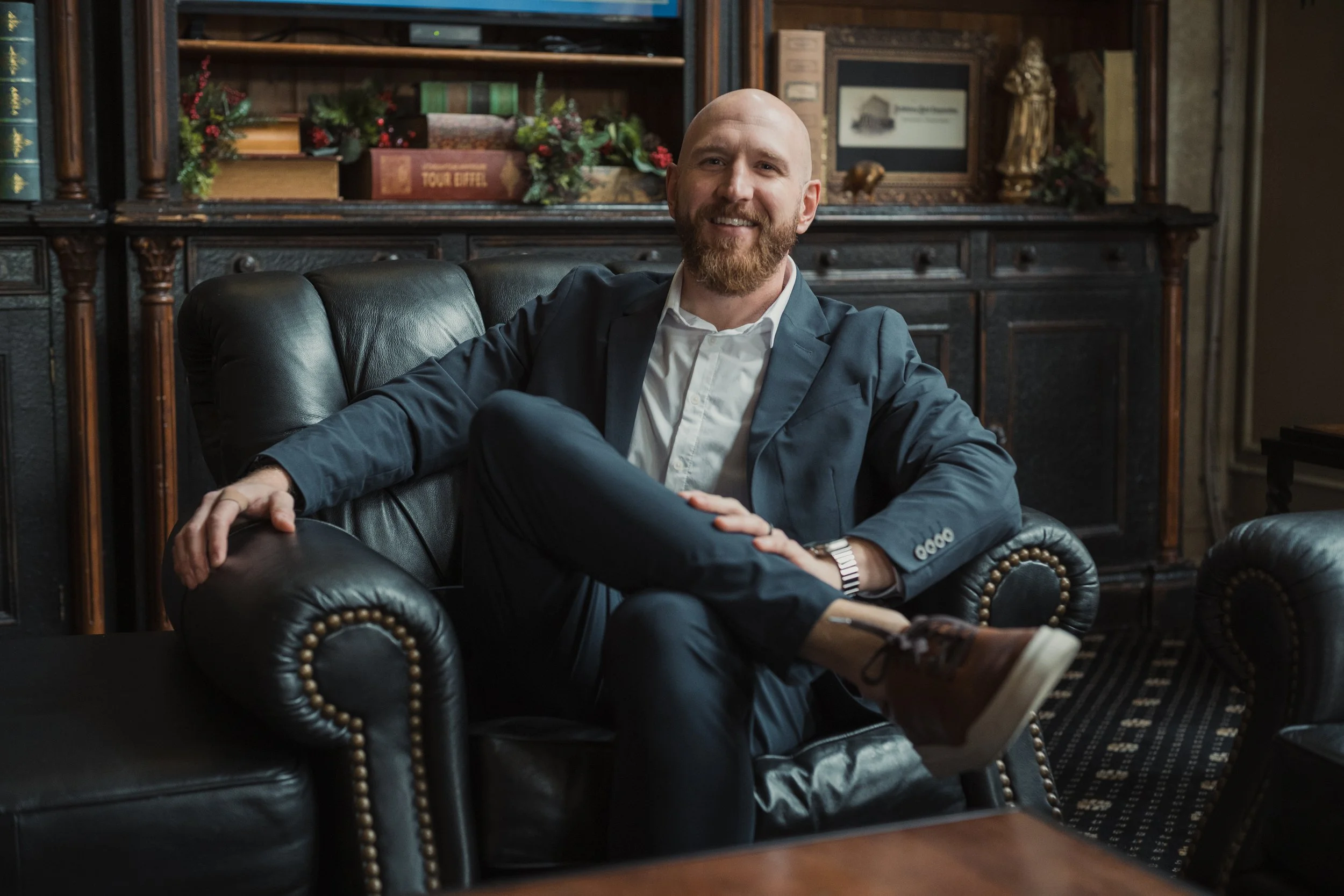A man with a beard is sitting on a black leather armchair in a room with dark wooden furniture and bookshelves, smiling and looking at the camera.
