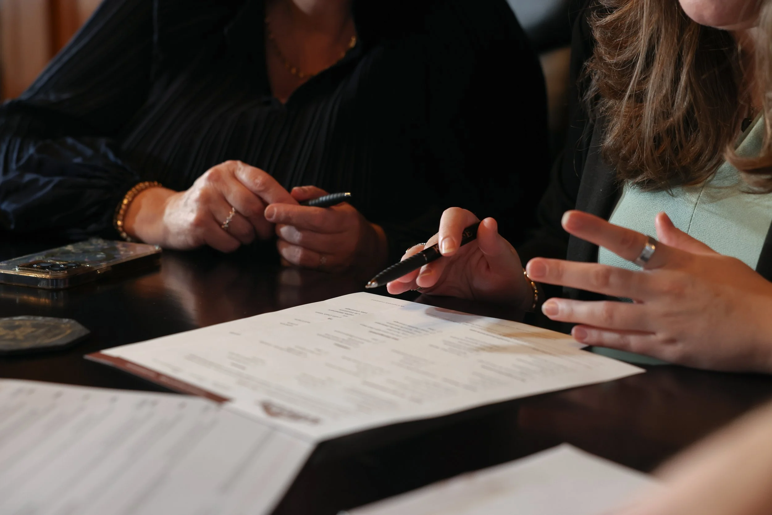 Two women sitting at a table, reviewing a document with pens in hand, near a smartphone and multiple papers.