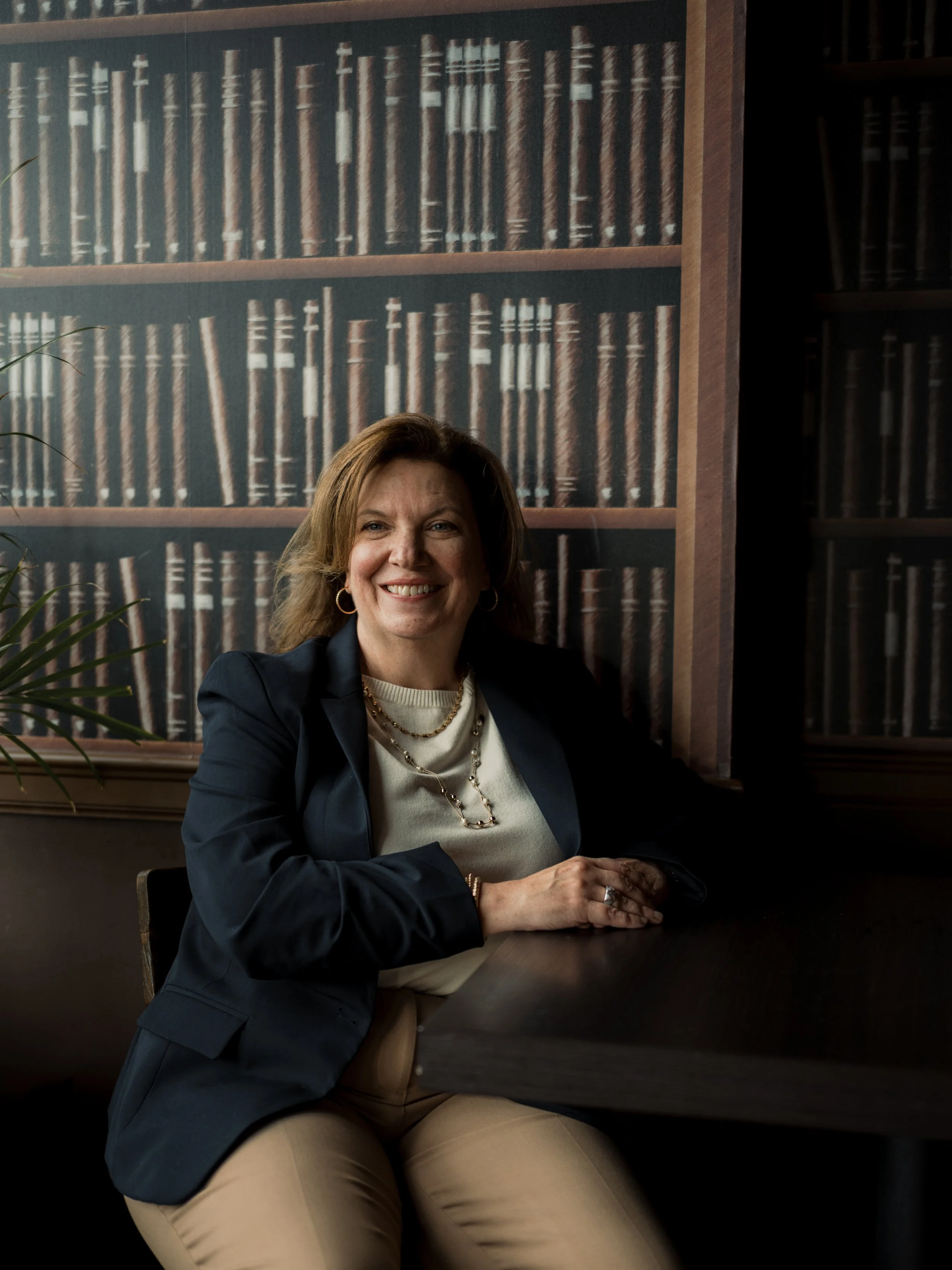 A woman with shoulder-length brown hair, wearing a dark blazer, cream top, and layered jewelry, sits at a dark wooden table in a room with a large bookshelf mural in the background and a potted plant partially visible on the left.