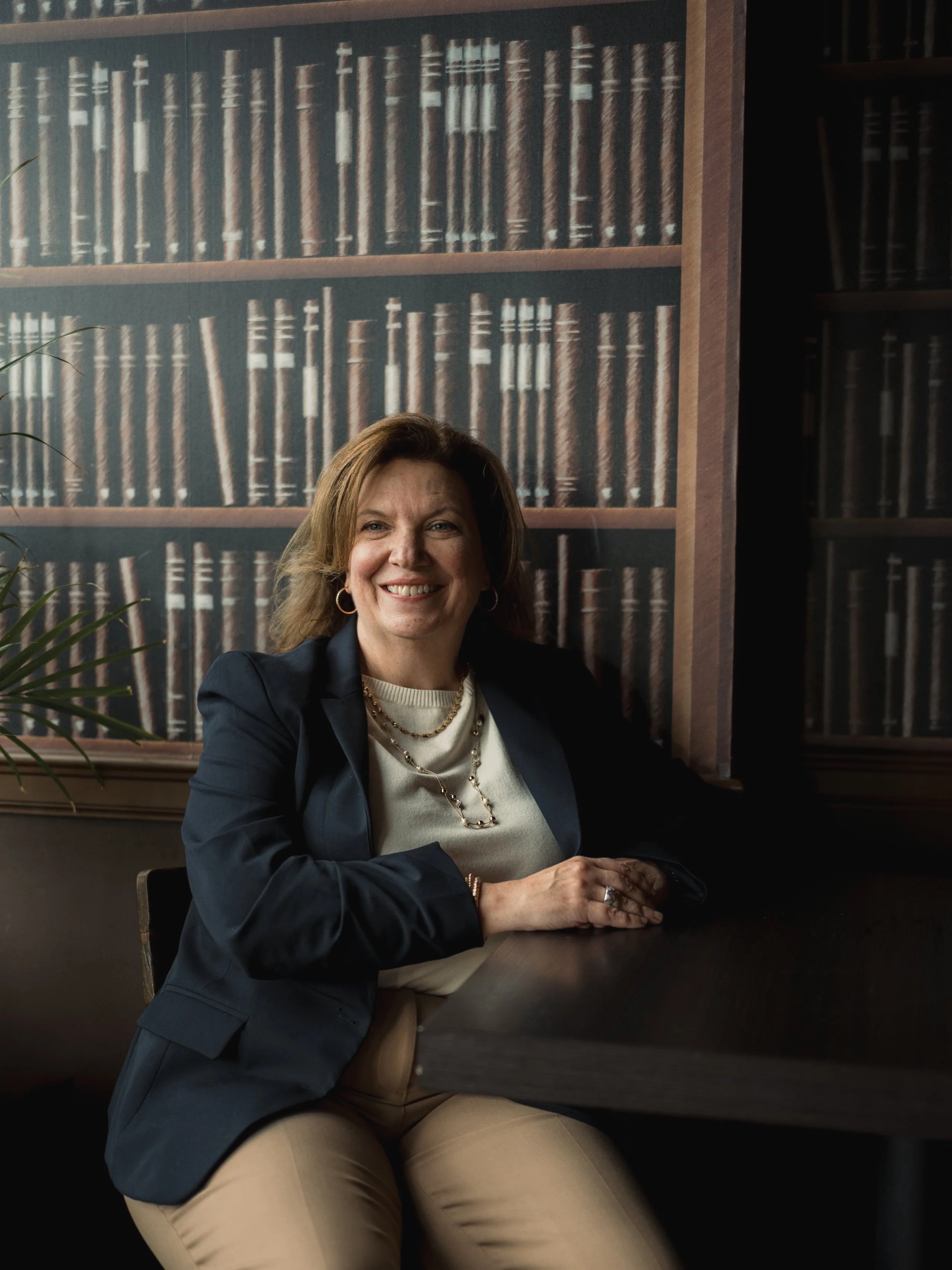 A woman sitting at a dark wooden table in front of a large mural of a bookshelf filled with books, smiling at the camera.