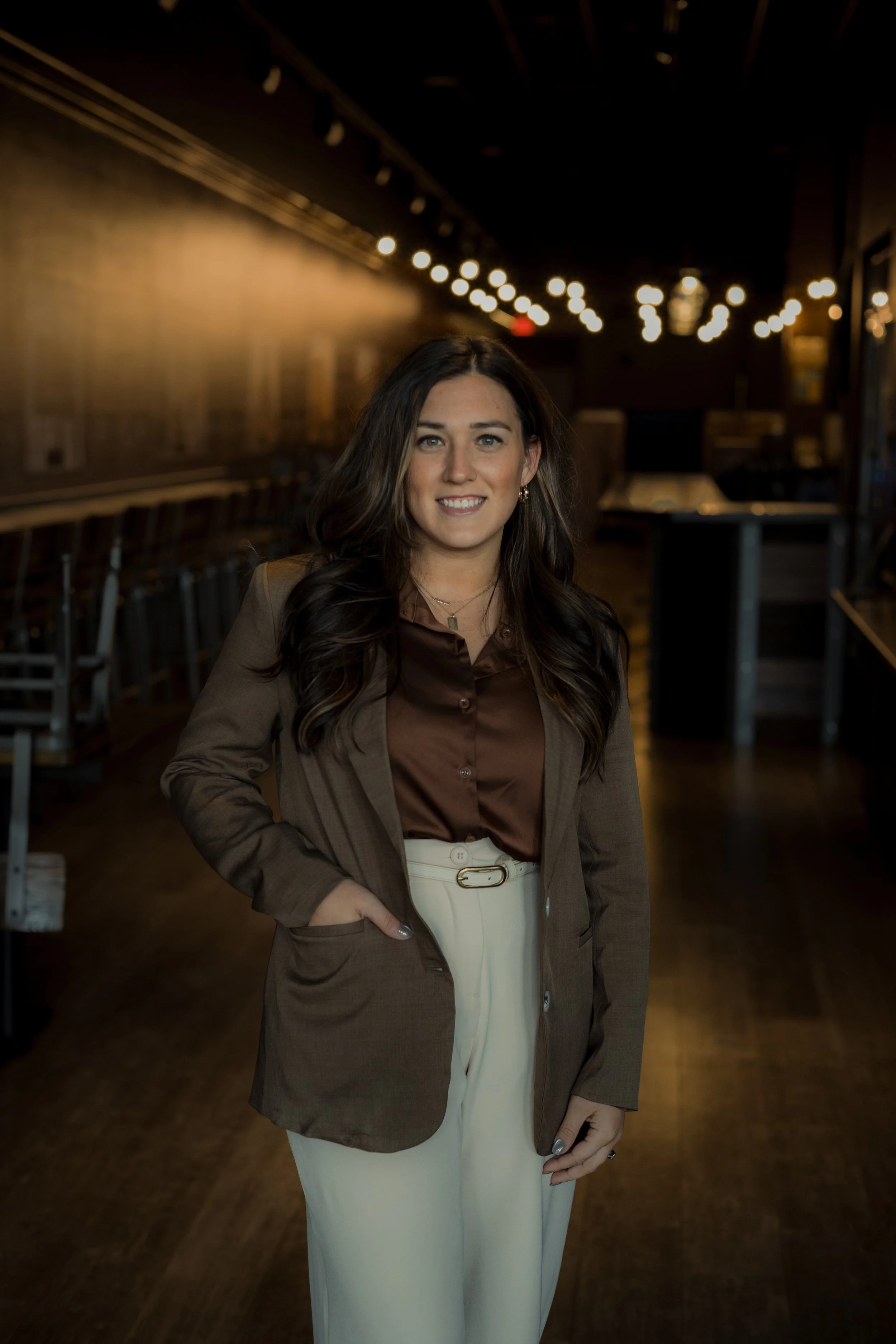 A woman with dark, wavy hair and a warm smile standing in a dimly lit room with string lights overhead, wearing a brown satin blouse, beige wide-leg pants, and a brown blazer.
