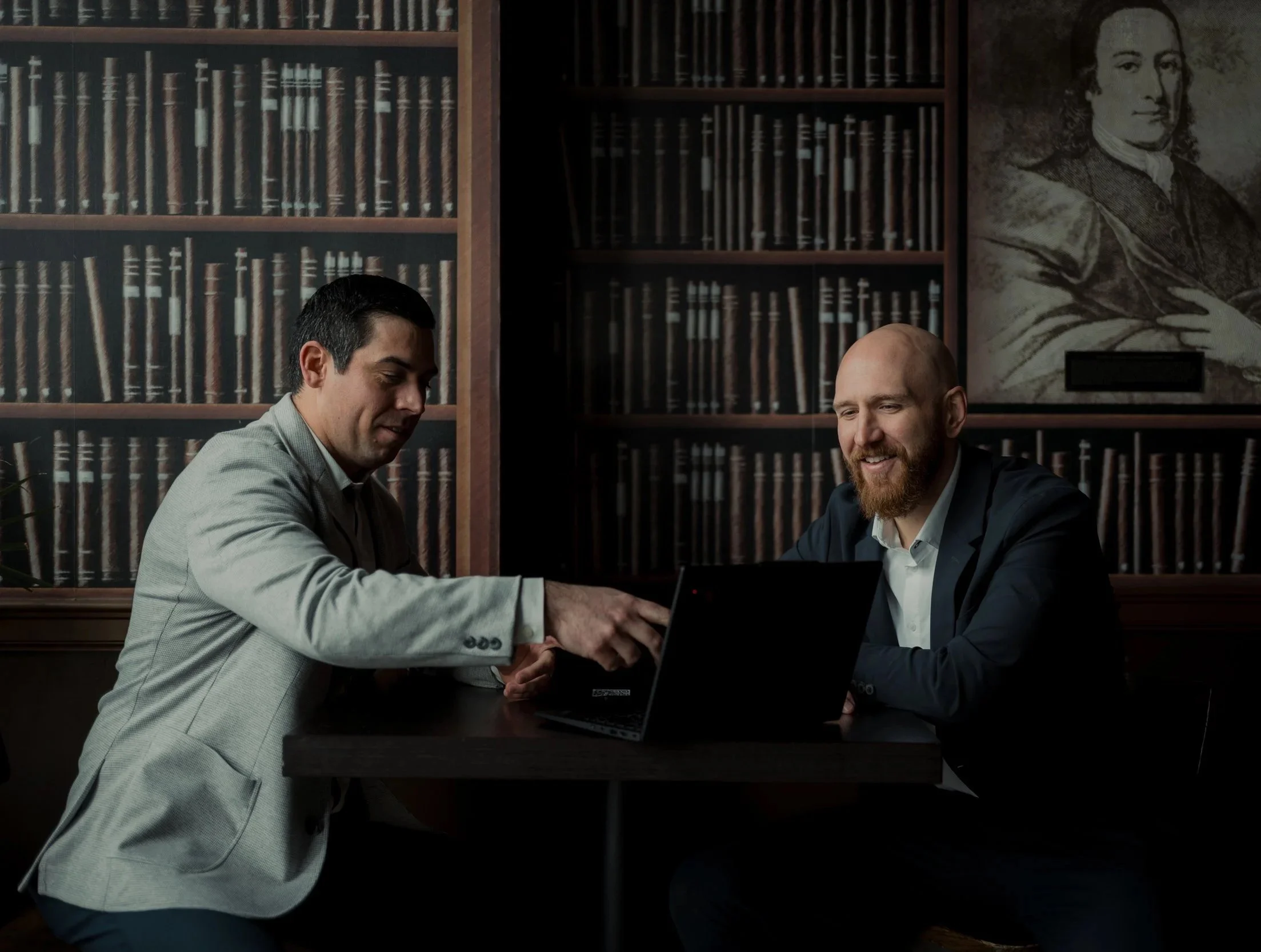 Two men in suits sitting at a table with laptops, exchanging documents in a library with bookshelves and a portrait of a historical figure in the background.