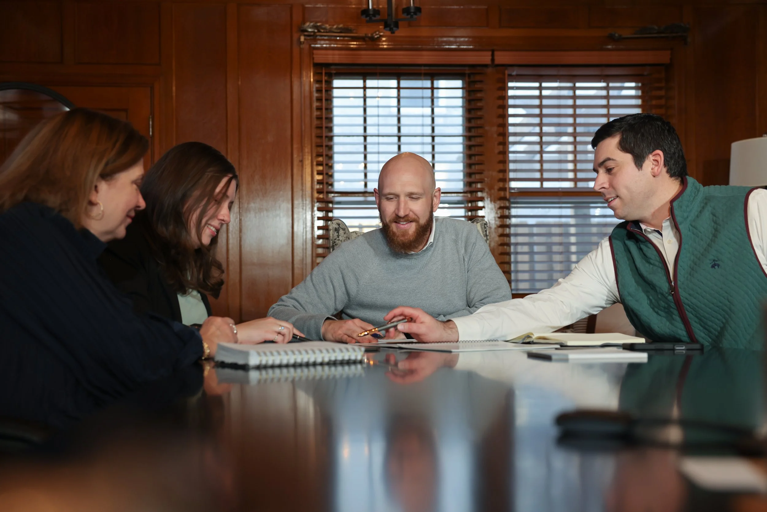 Four people sitting around a wooden table in a room with wooden walls and blinds, engaging in a discussion. A man with a beard and a gray sweatshirt is smiling, while the others are looking at documents and a phone.