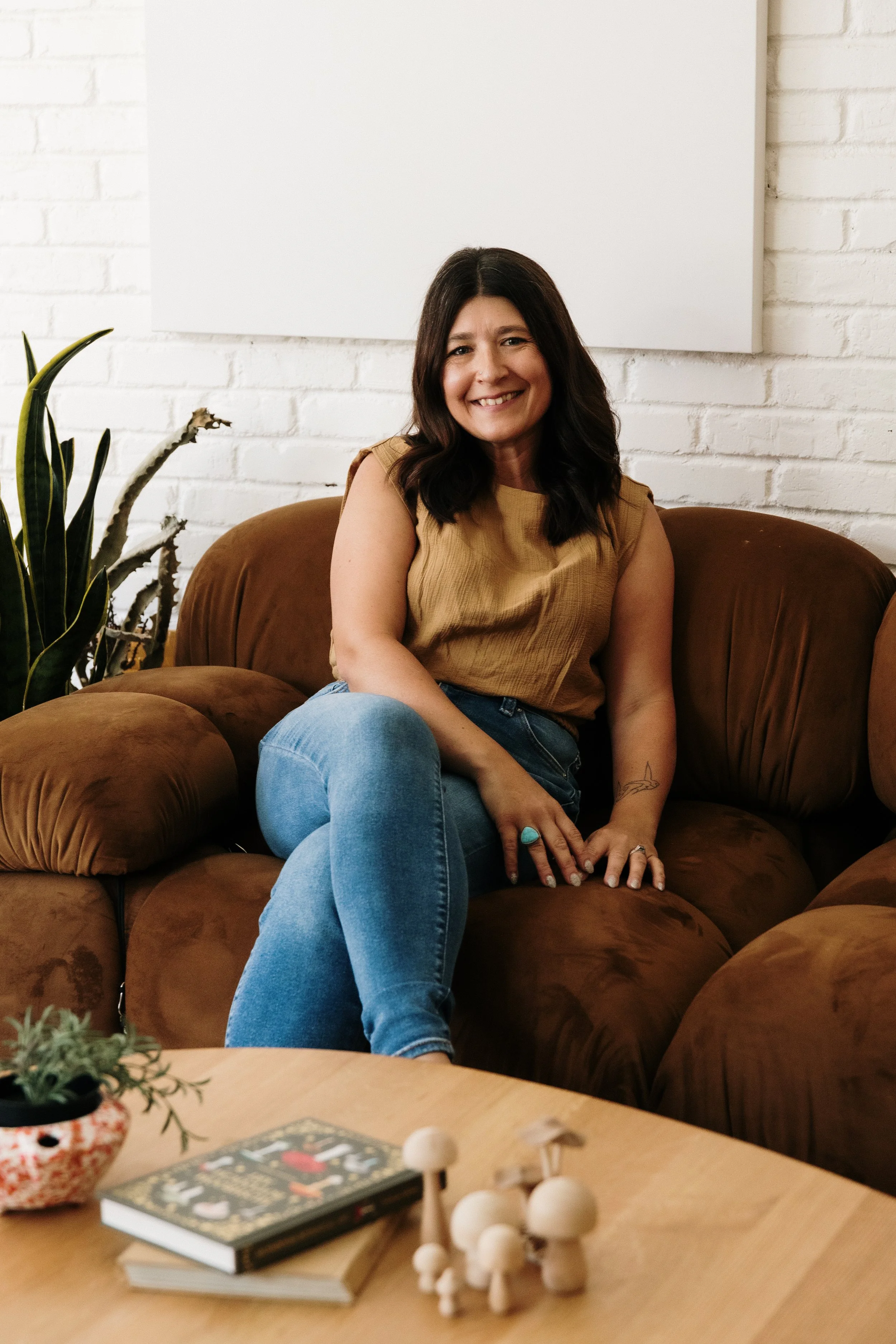 A woman with dark hair, wearing a sleeveless camel-colored top and blue jeans, sits on a brown suede sofa, smiling at the camera in a cozy living room with white brick walls, a plant, a coffee table with books, a small decorative mushroom sculpture, and a tattoo on her left arm.