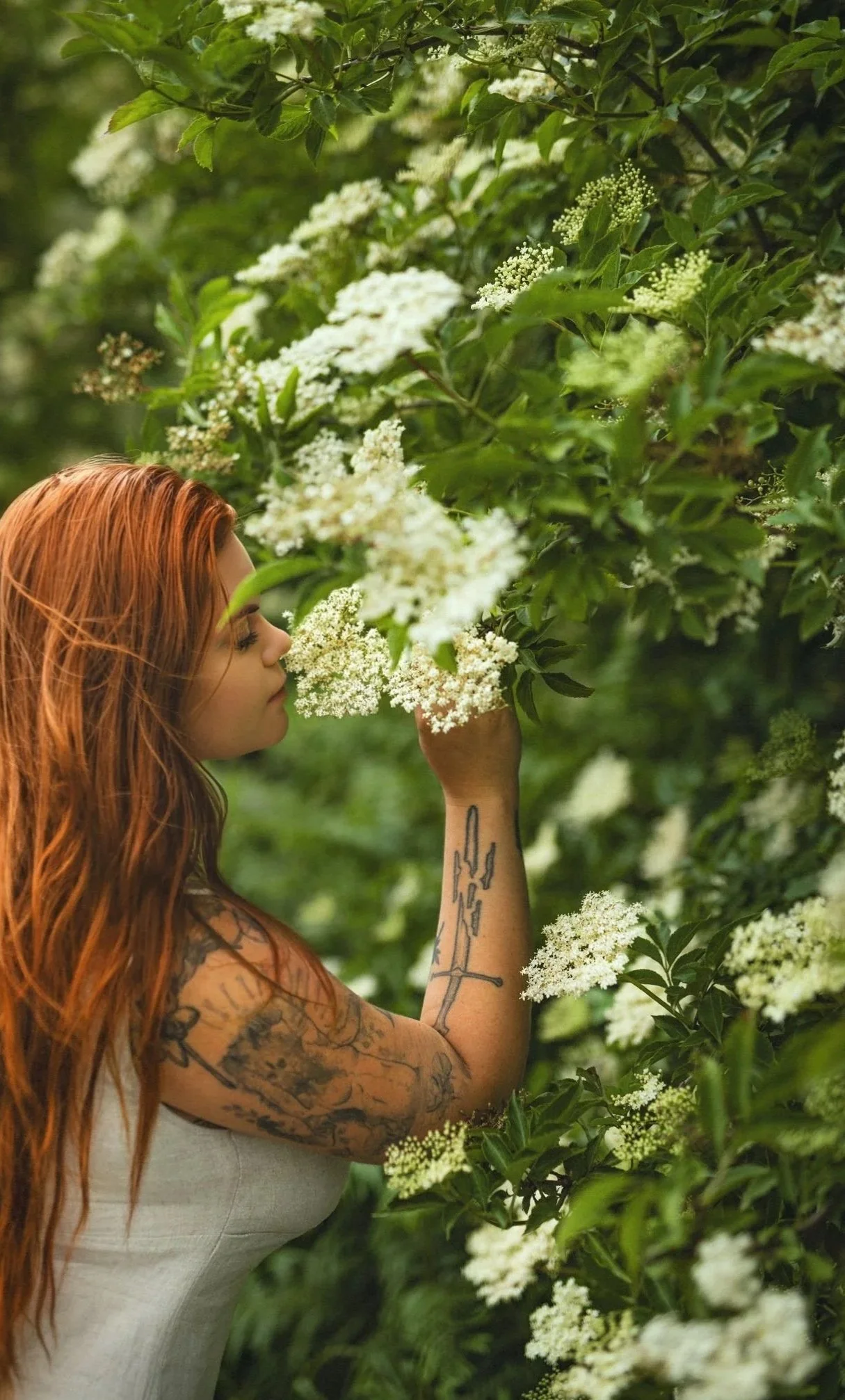 A woman with long red hair and tattoos on her arm is smelling white flowers on a bush in a lush green garden.