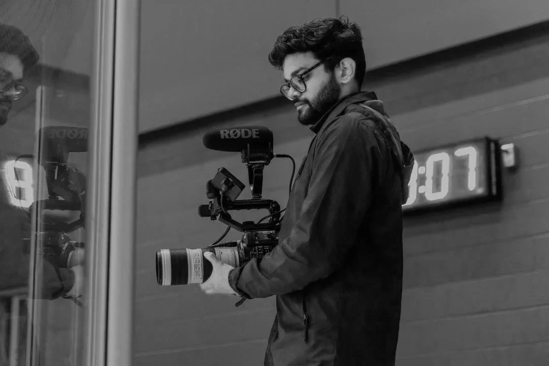 A man with glasses and a beard holding a professional video camera, facing a mirror or glass surface, with a digital clock showing 1:07 in the background.