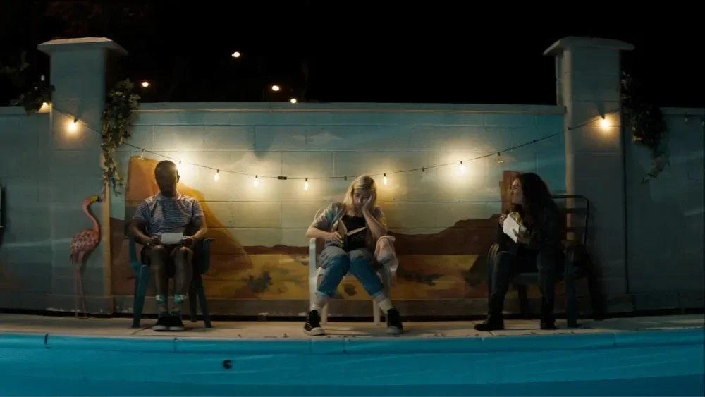 Three women sitting on chairs in front of a painted scenic backdrop with string lights, near a swimming pool at night.