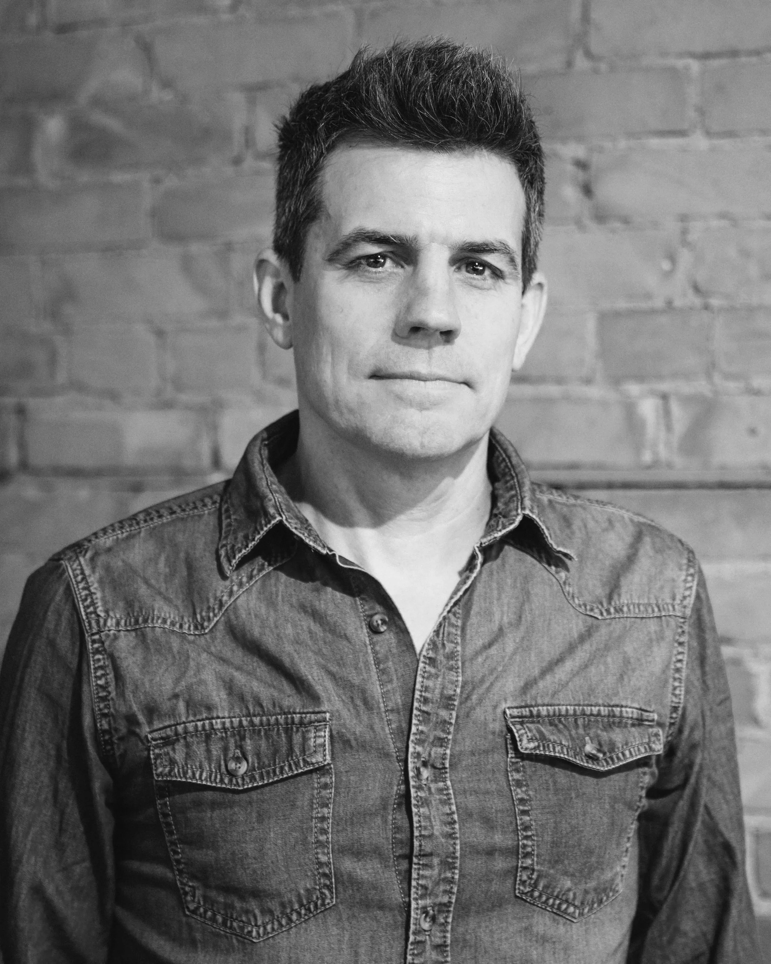 Black and white portrait of a man with short dark hair, wearing a denim shirt, standing against a brick wall background.