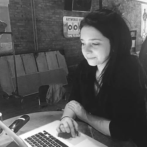 Young woman sitting at a table using a laptop, with wooden walls and artwork in the background.