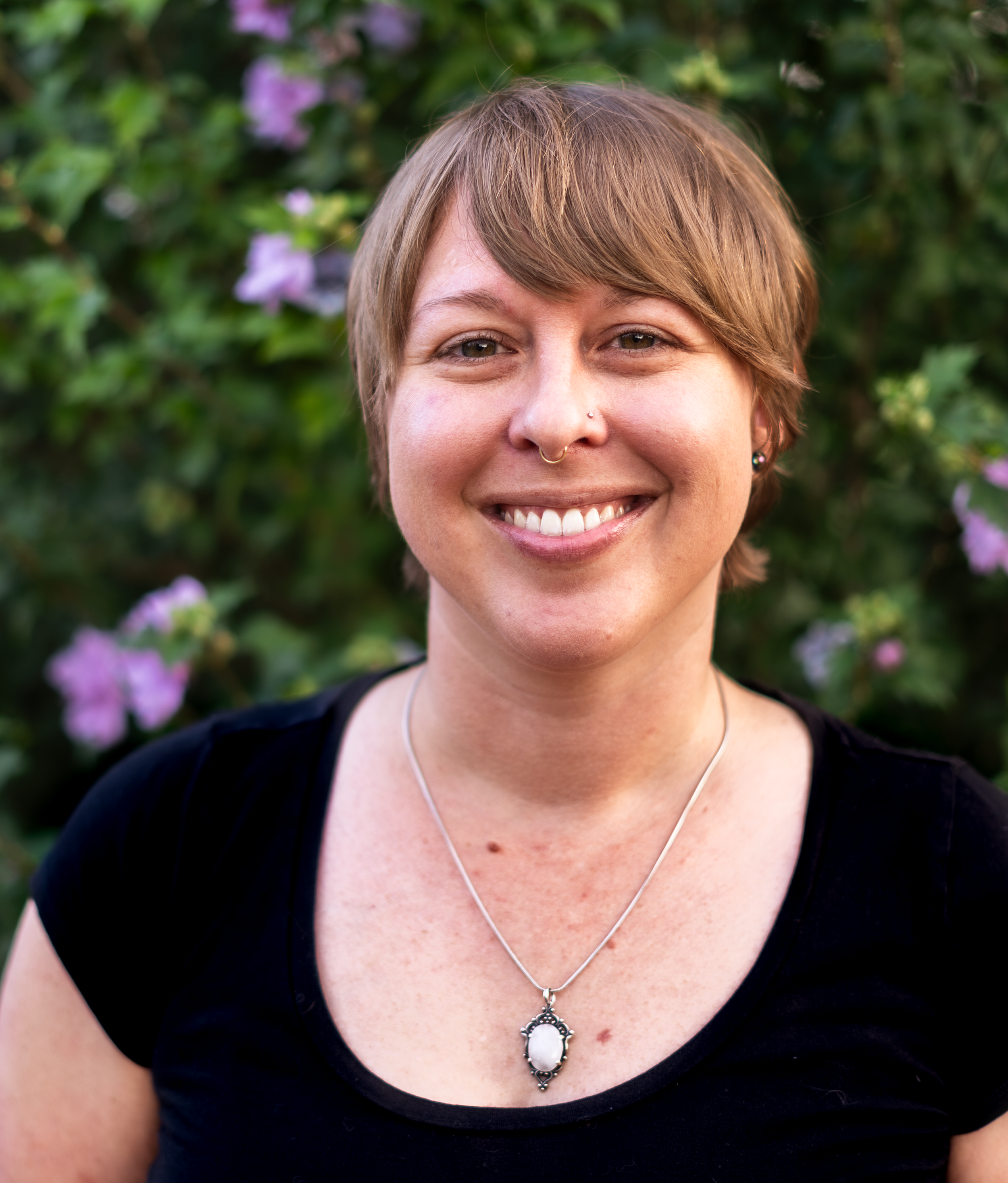 A therapist with short light brown hair smiles gently in an outdoor setting, surrounded by soft green foliage and purple blooms. Their expression is warm and steady, conveying a sense of grounded presence and soulful connection.