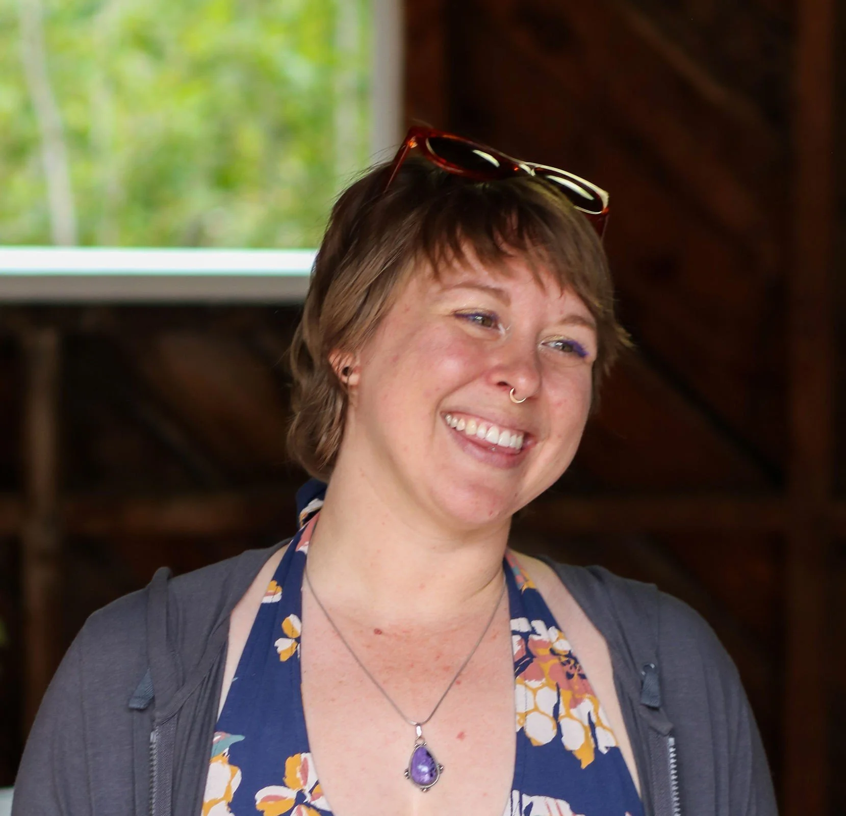 Caroline Rapp smiling with short brown hair, sunglasses on their head, wearing a floral dress and a purple gemstone necklace, standing indoors with a wooden wall and window showing greenery outside.