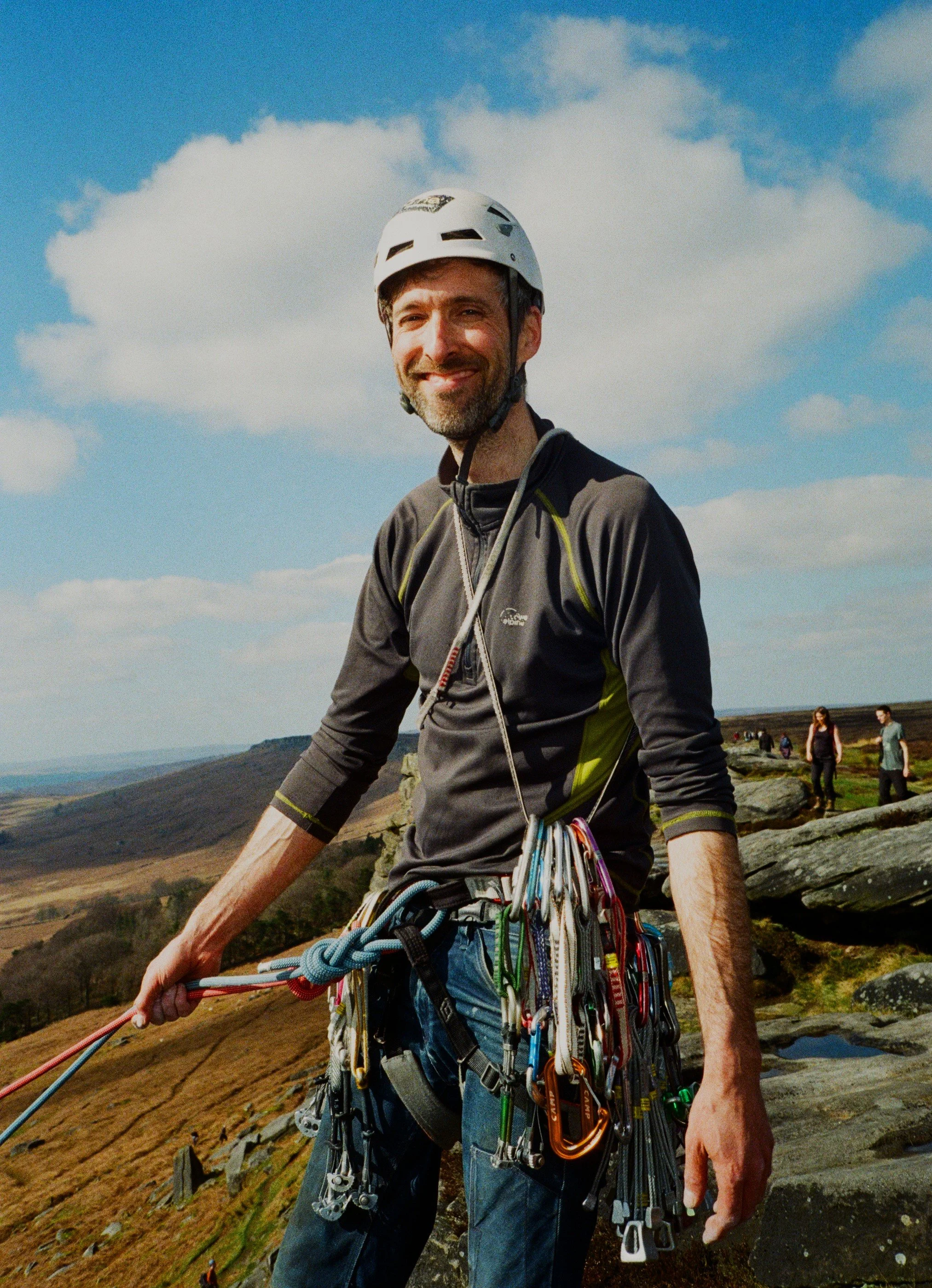 A 35mm film portrait of a man wearing trad climbing gear and a white helmet stands at the top of Stanage rocks. He has several quick draws, cams and climbing equipment hanging off him.