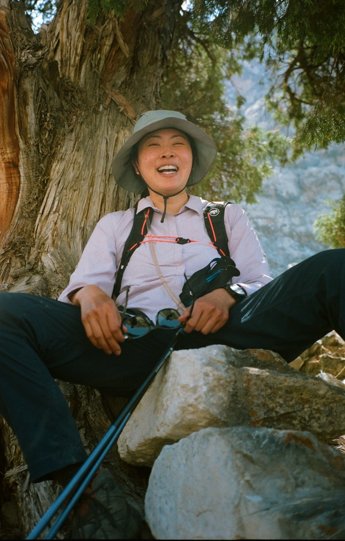 A woman wears hiking gear and has taken her sunglasses off as she rests by a tree in the Fann Mountains, Tajikistan. 35mm film photography.