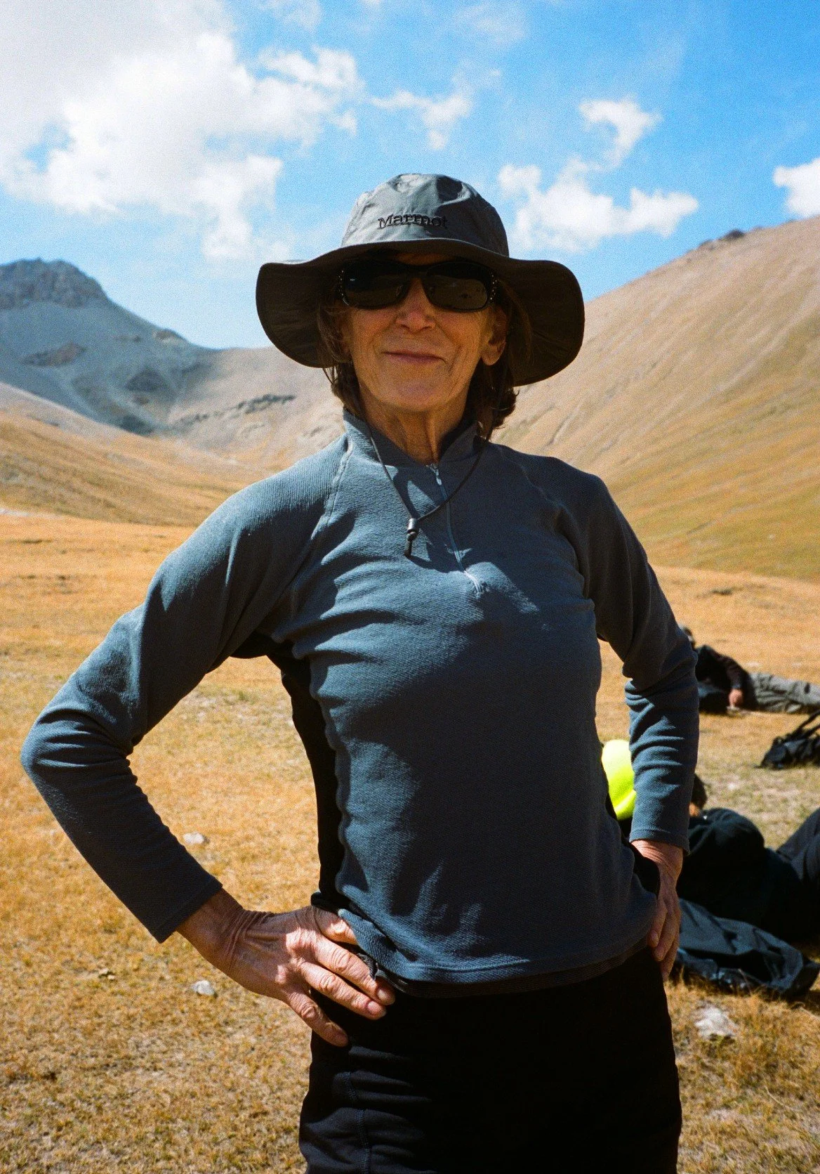 In the mountains of Kyrgyzstan, Central Asia, a woman wears black hiking clothing and a black Marmot sun hat. She has her hands on her hips and smiles at the camera.