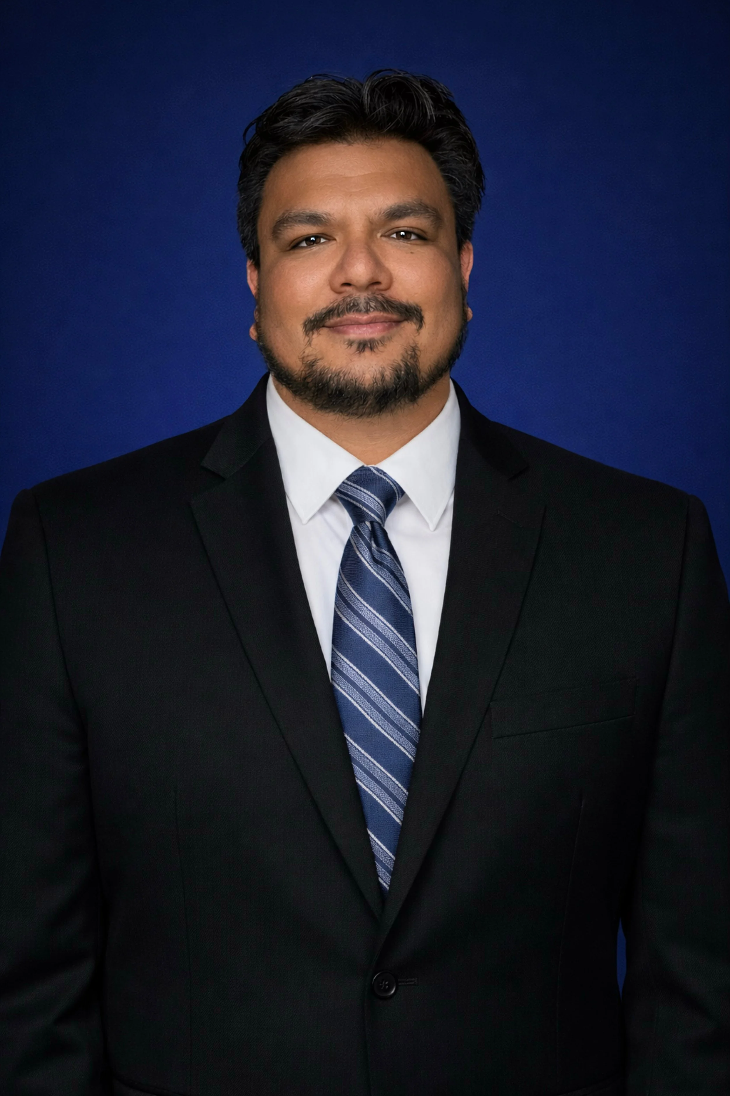 Professional portrait of a man in a black suit, white shirt, and striped blue tie, standing against a dark blue background.