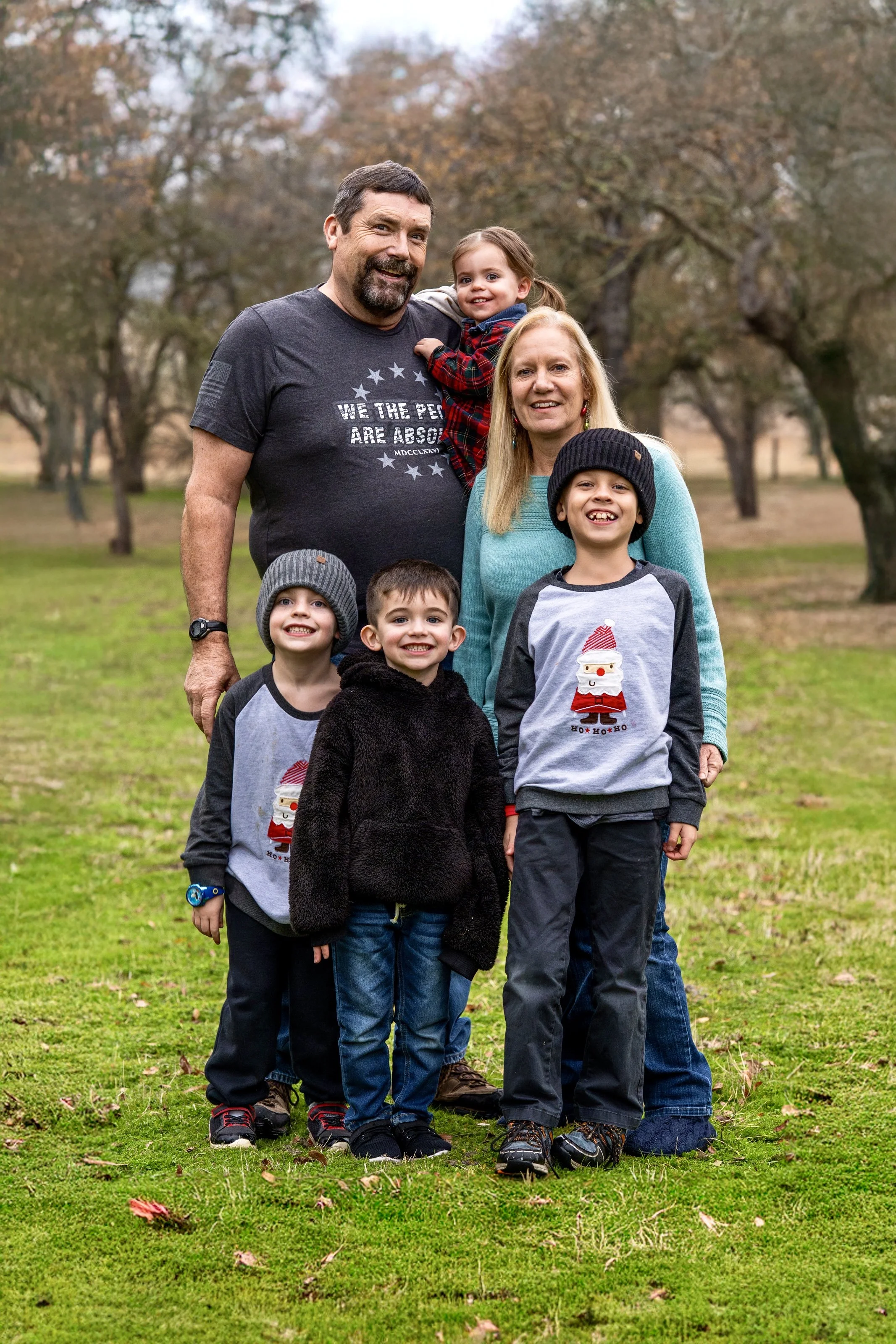 A family of six standing outdoors on a grassy field, smiling at the camera during autumn, with trees in the background. The family includes two adults and four children, some wearing holiday-themed clothing.