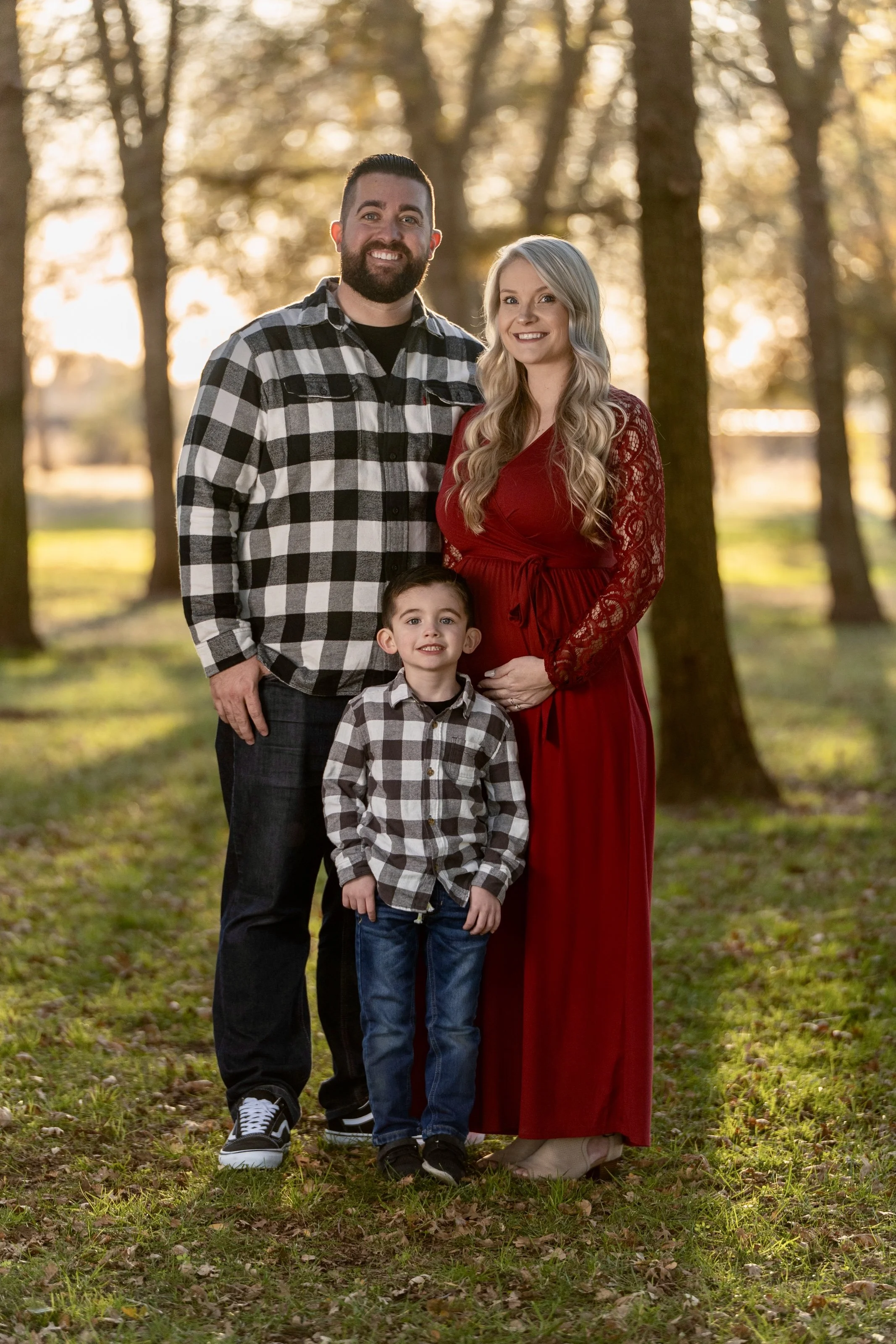 A family of three standing outdoors in a park during autumn, with trees and sunlight in the background. The man has dark hair and a beard, wearing a black and white plaid shirt and black pants. The woman has long blonde hair, wearing a long red dress