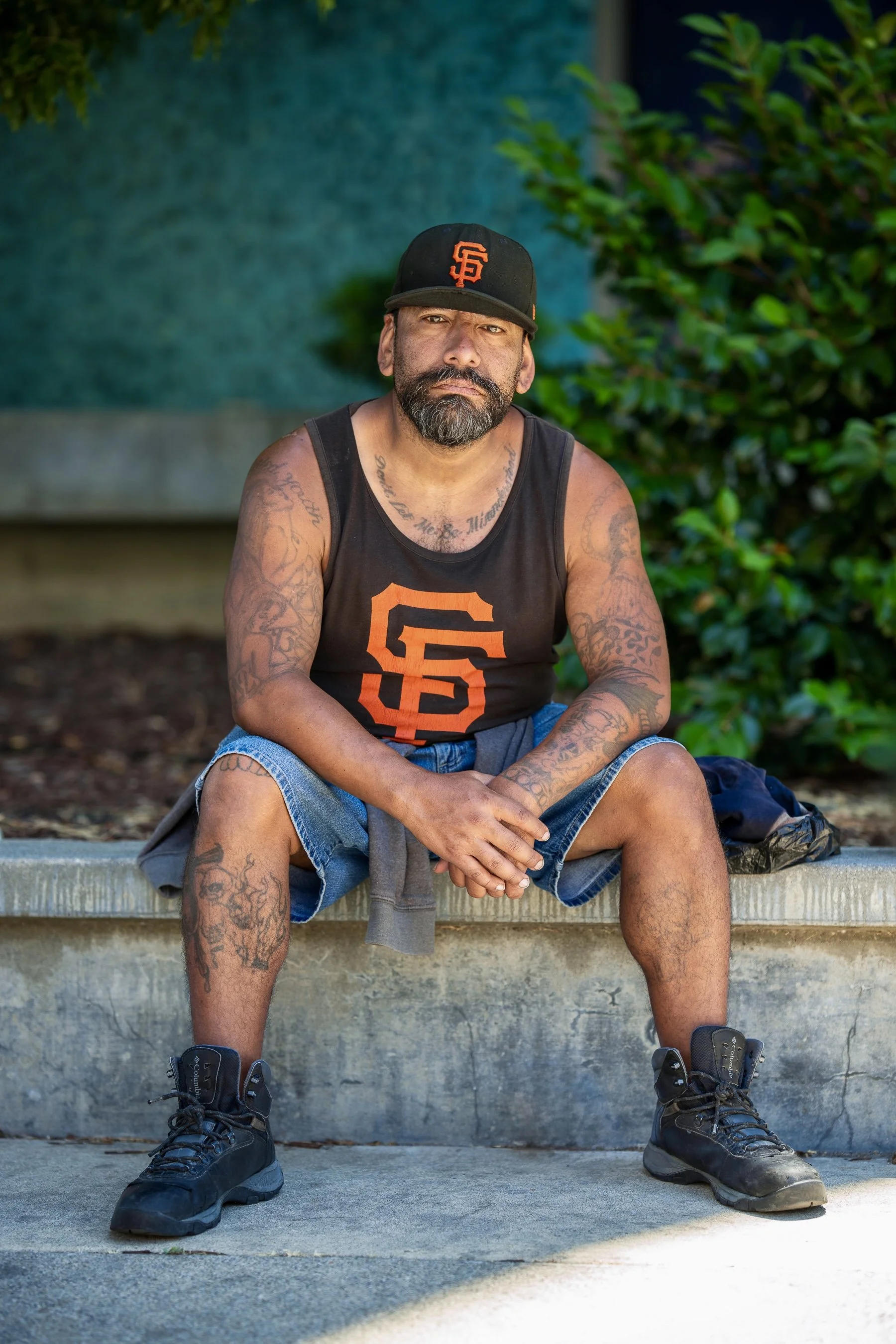 A man with tattoos, a beard, and a mustache sitting on a concrete ledge outdoors, wearing a black San Francisco Giants cap, black sleeveless shirt with Giants logo, denim shorts, and black hiking boots.