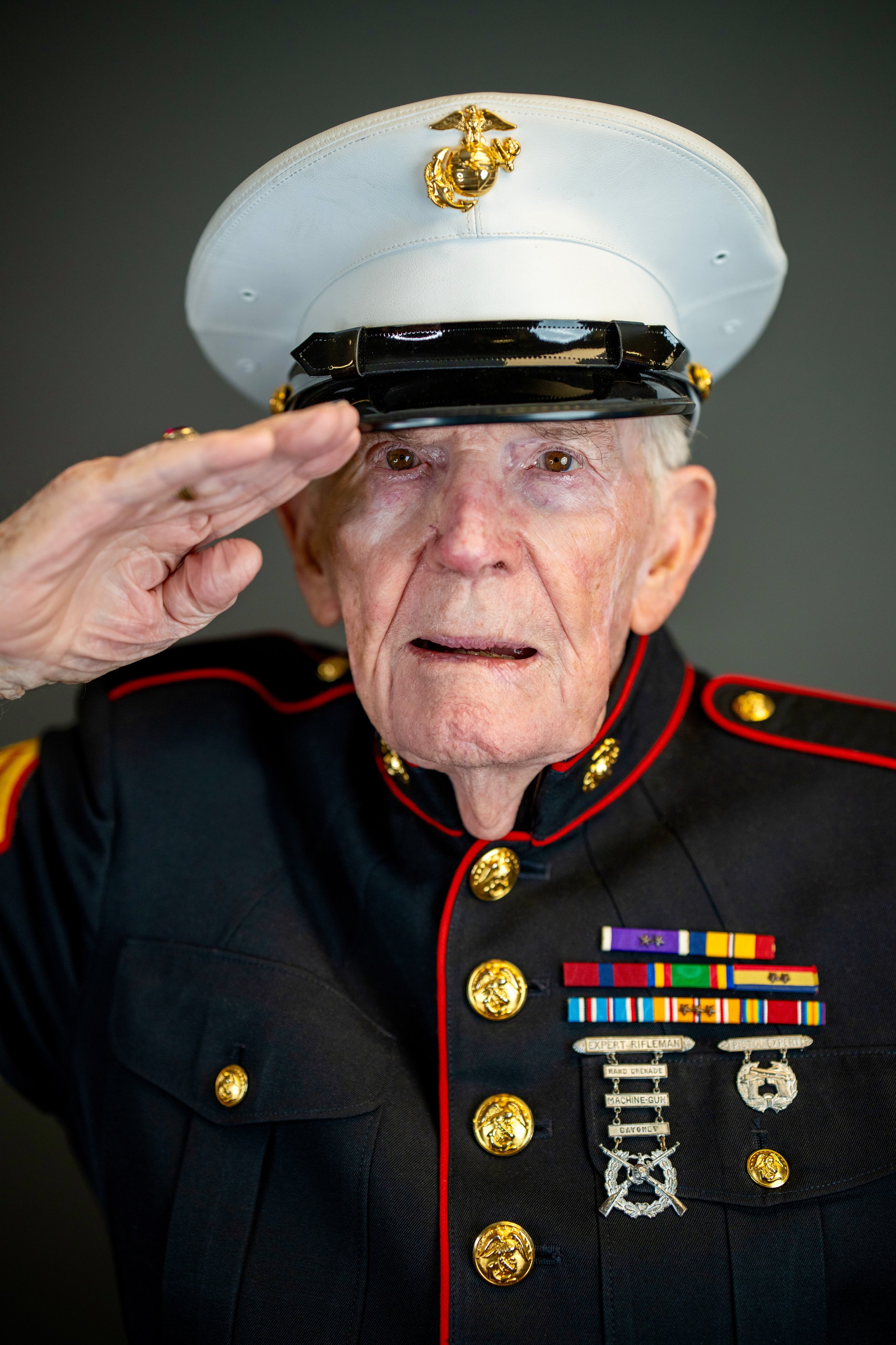 An elderly Marine veteran salutes while wearing his military uniform and hat with the Marine Corps emblem. He has several medals and ribbons on his chest.