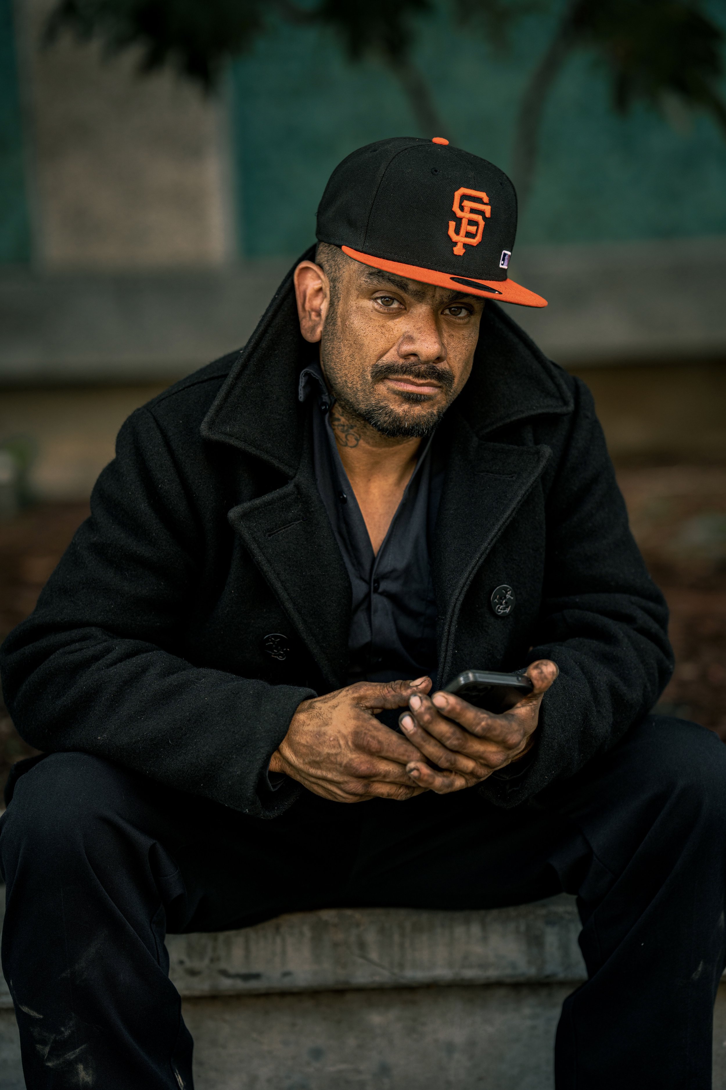 A man sitting outdoors on a bench, wearing a black coat and a black and orange baseball cap with the San Francisco Giants logo, holding a smartphone in his hands and looking at the camera.
