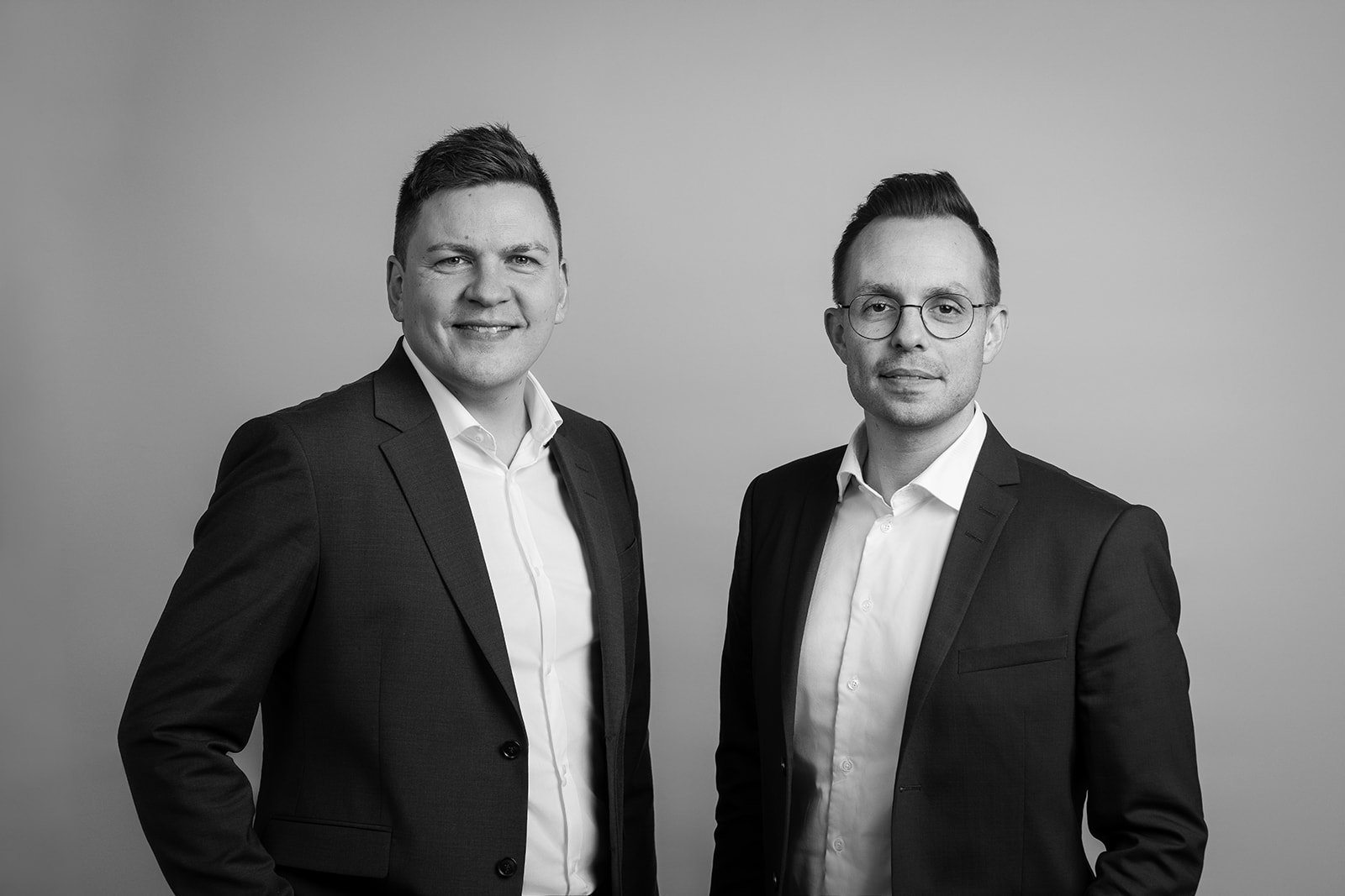 To mænd i forretningsdragter står foran en neutral baggrund, fotograferet i sort og hvid.
