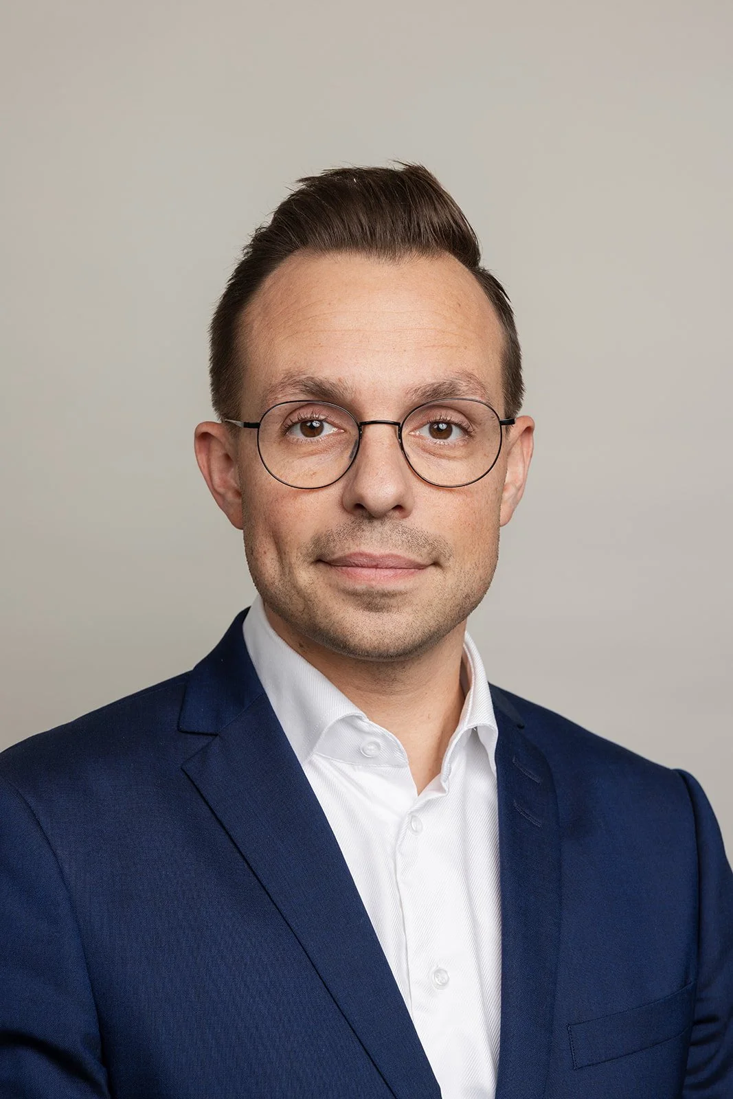 Portræt af en mand med brunt hår, briller, iklædt en hvid skjorte og en mørkeblå blazer, foran en neutral baggrund.