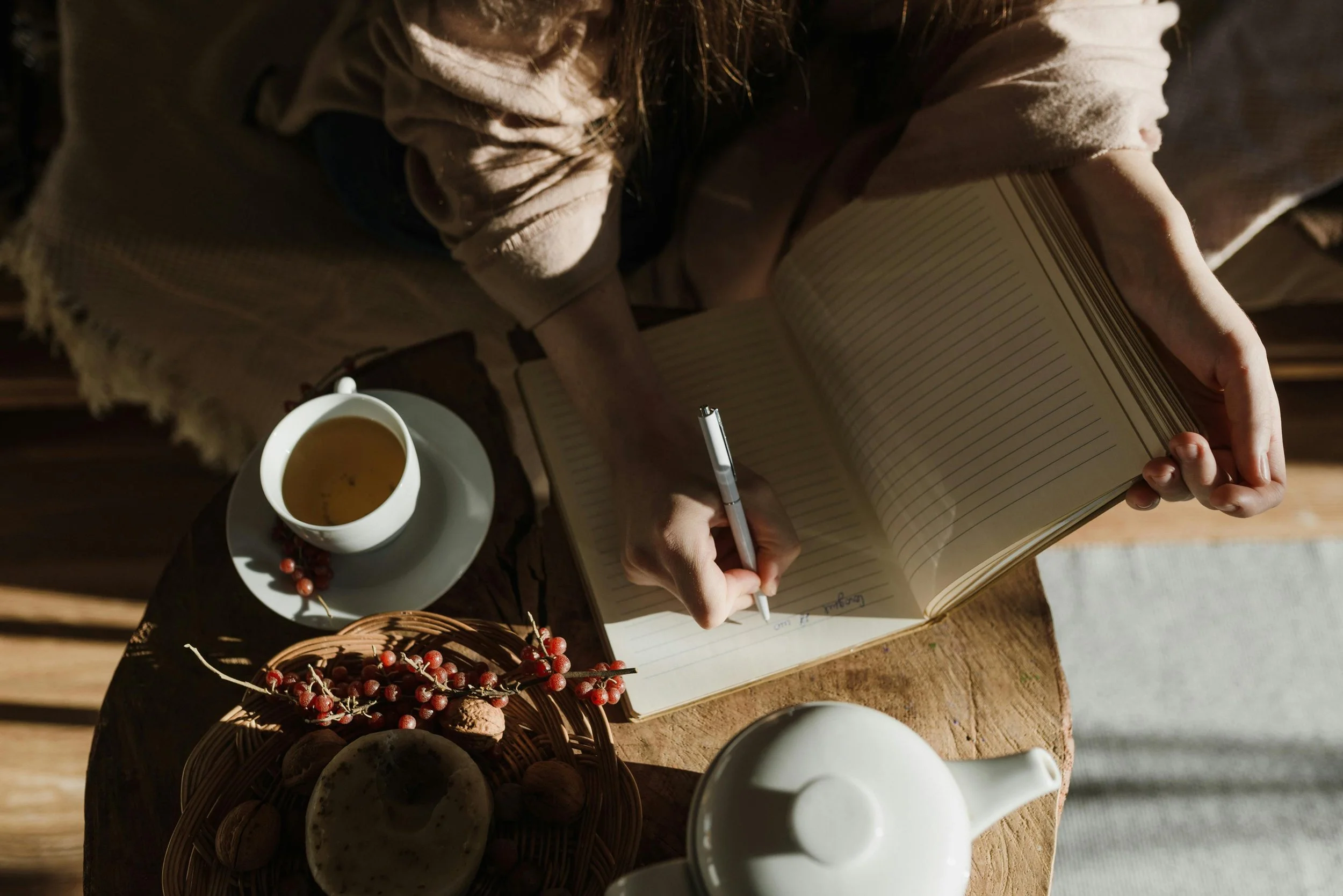 A person journaling in a notebook with a cup of tea or coffee, a basket of cookies, and a cluster of red berries on the table. The scene is lit with warm, natural lighting.