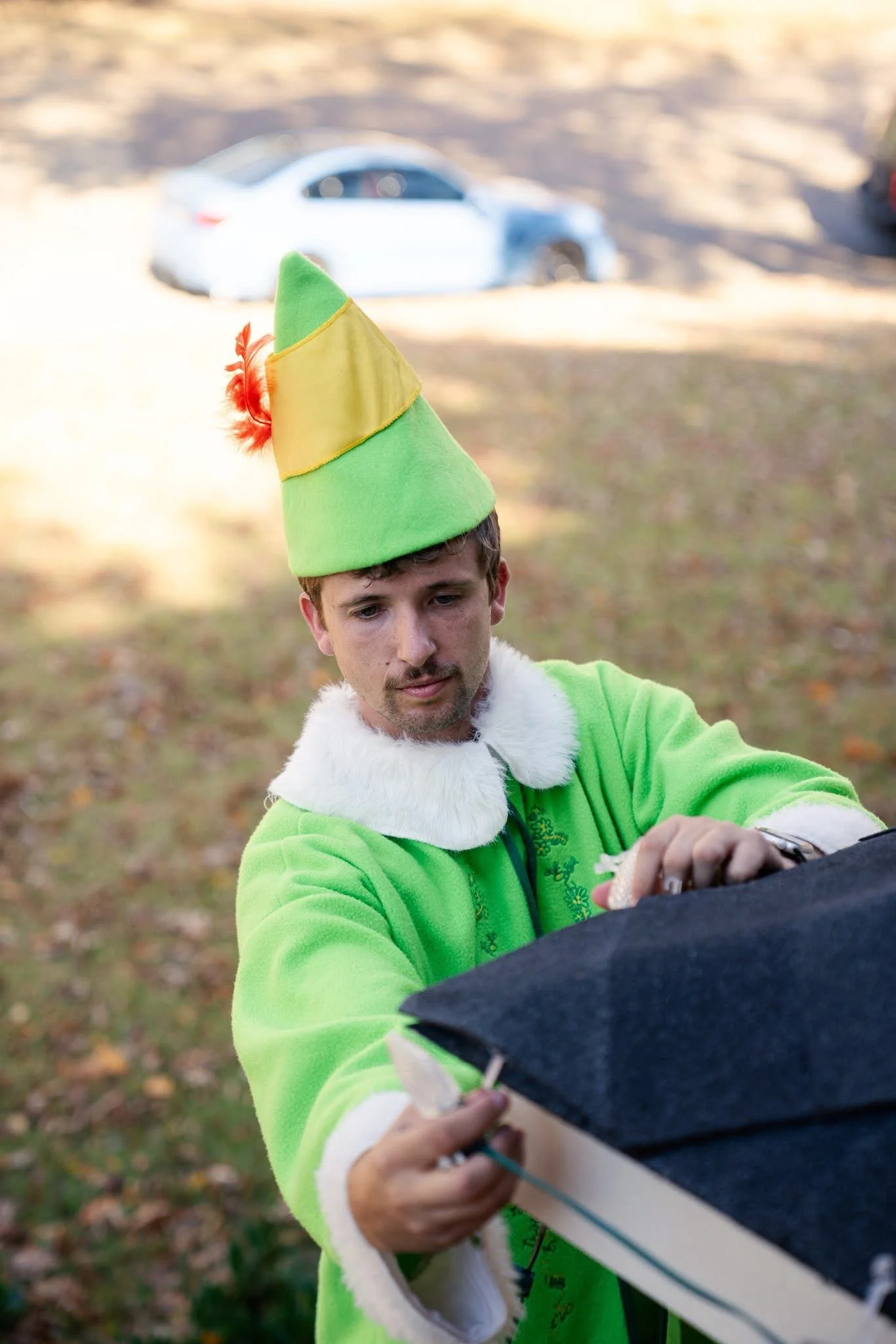A man dressed in a bright green elf costume with a pointed hat that has orange feathers, adjusting a black box outdoors during daytime.