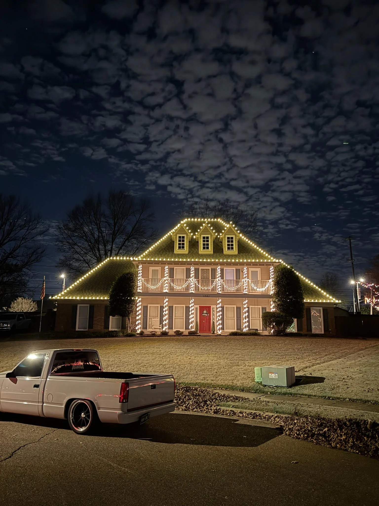 A house decorated with Christmas lights at night, featuring a pitched roof, small dormer windows, and a red front door, with a pickup truck parked in front and a partly cloudy sky overhead.