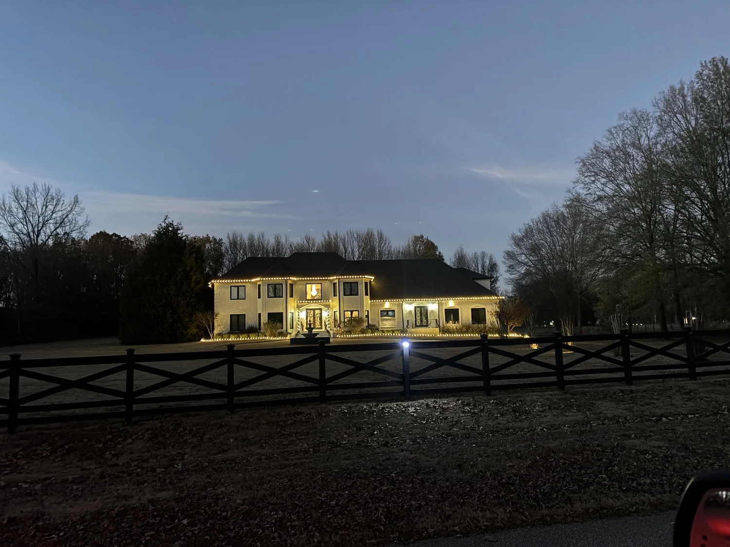 Country home with holiday lighting seen across a fenced yard
