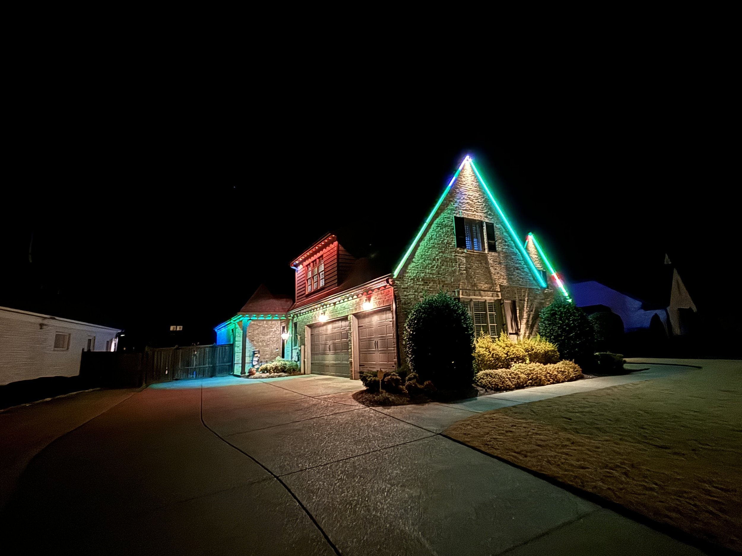 Night view of a house decorated with colorful Christmas lights, including green, red, and blue, outlining the roof and windows.