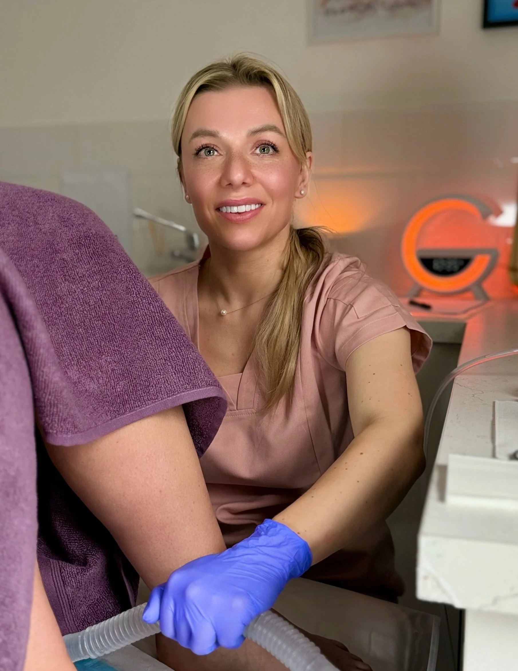 A female healthcare professional with blonde hair smiling during a medical procedure with a patient's leg visible, wearing purple gloves, in a clinical setting with medical equipment and an orange Glooworm device in the background.