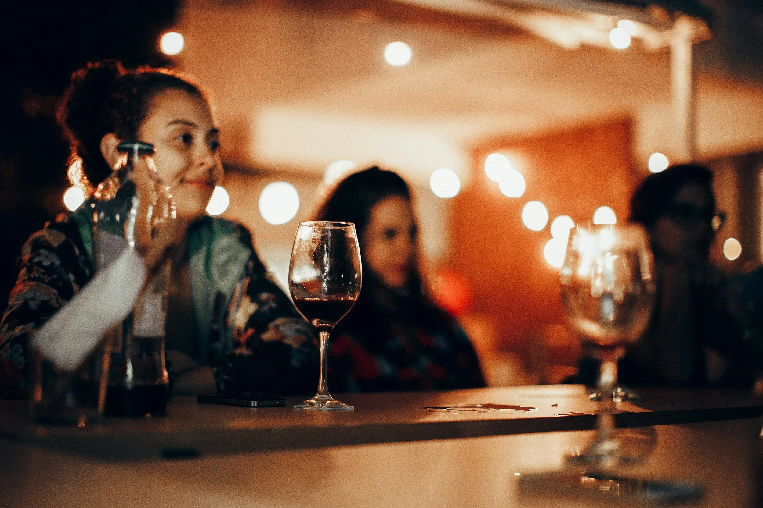 A woman and two other people sitting at a table in a dimly lit restaurant or bar, with a glass of red wine in the foreground, celebrating or enjoying a social moment.
