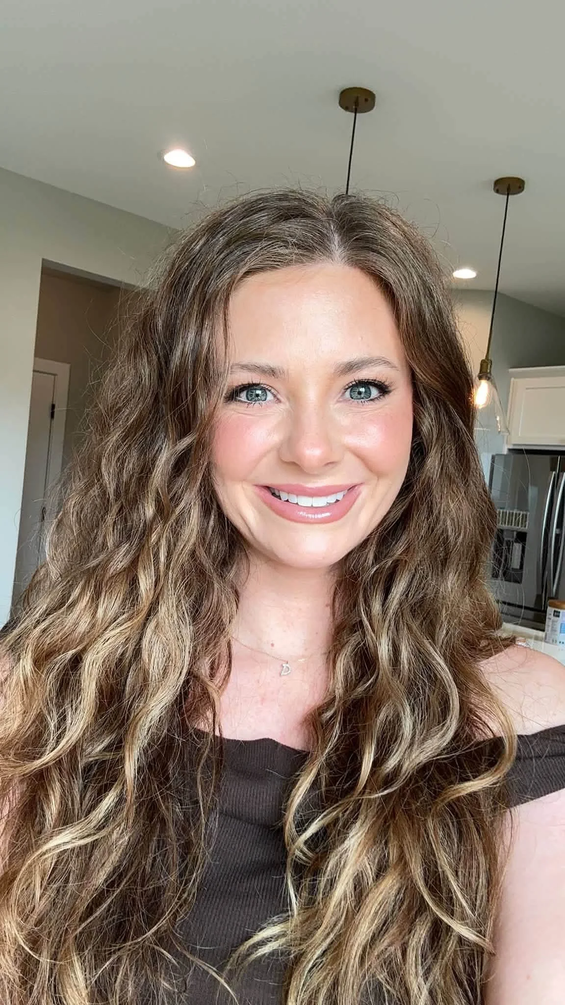 A woman with long, curly light brown hair and blue eyes smiling in a kitchen with hanging pendant lights.