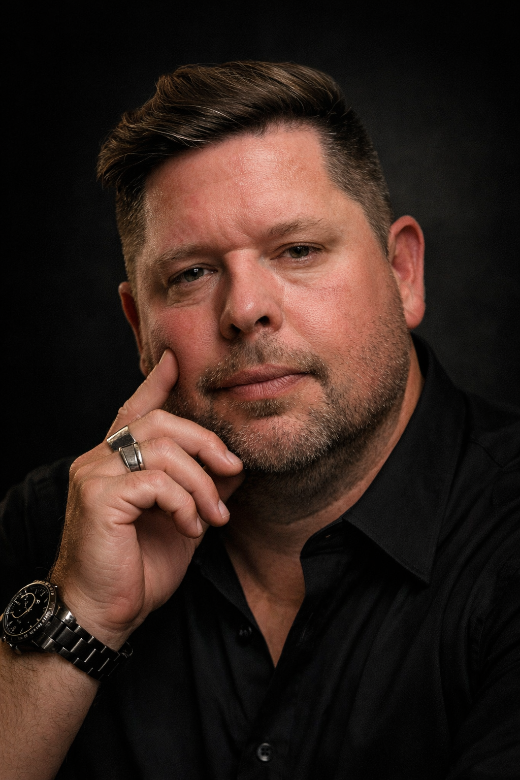 A man with short, dark, styled hair and a beard, smiling, wearing a patterned shirt with floral designs, standing outdoors with palm trees and a clear, light sky in the background.