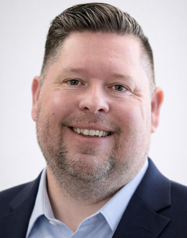 A man with short, dark, styled hair and a beard, smiling, wearing a patterned shirt with floral designs, standing outdoors with palm trees and a clear, light sky in the background.
