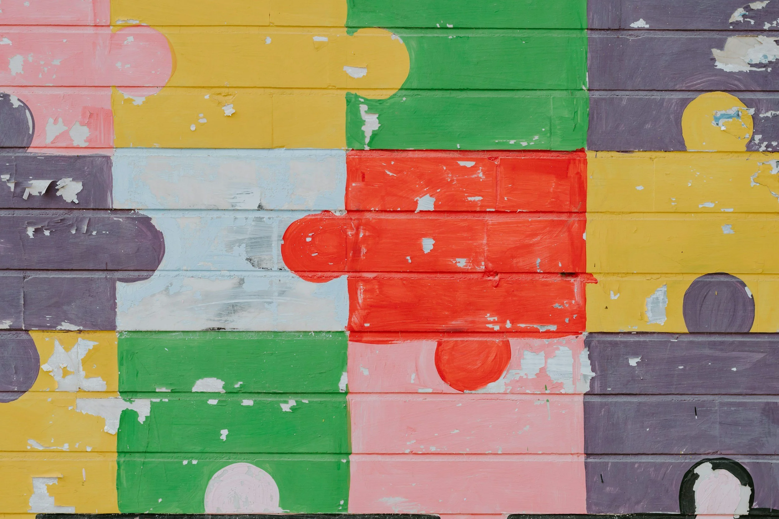 Colorful abstract mural painted on wooden planks with patches of peeling paint, featuring a jigsaw puzzle design in pink, yellow, green, purple, white, and black.