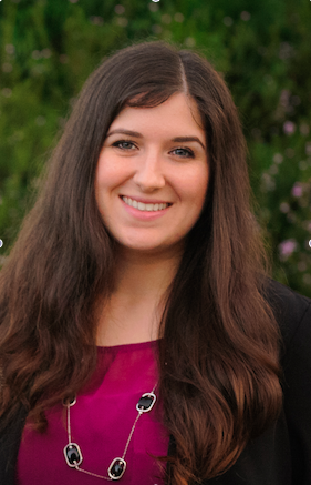 A young woman with long brown hair, wearing a black blazer, dark pink blouse, and necklace, smiling outdoors with greenery and purple flowers in the background. She is Dr. Micol Gonella the owner of Olive Trees Therapy.