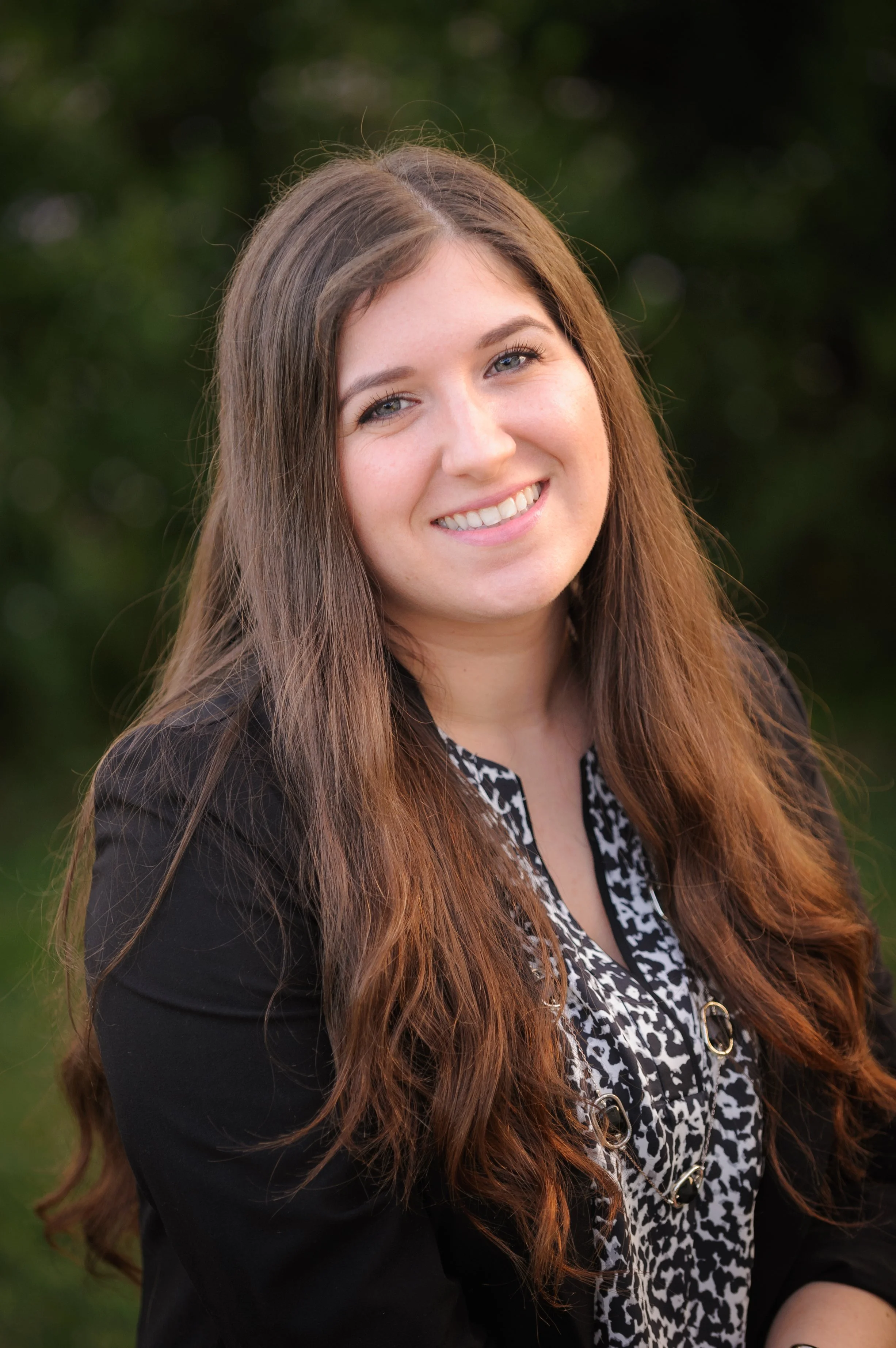 A young woman wearing a black blazer and black and white blouse, with long brown hair and blue eyes smiling for a photo outdoors with greenery in the background. She is Dr. Micol Gonella the owner of Olive Trees Therapy.