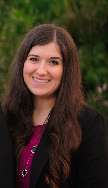 A photo of a young woman with long brown hair smiling outdoors with green foliage in the background. She is Dr. Micol Gonella the owner of Olive Trees Therapy.