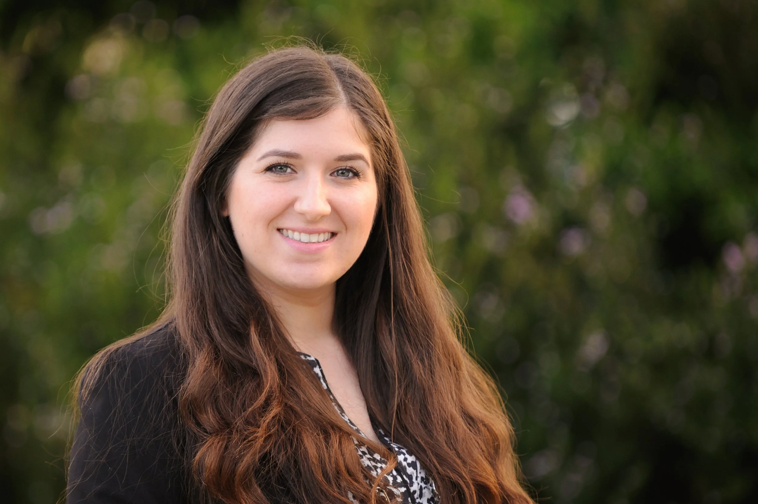 A young woman with long brown hair, wearing a black blazer and patterned top, smiling outdoors with greenery in the background. She is Dr. Micol Gonella the owner of Olive Trees Therapy.